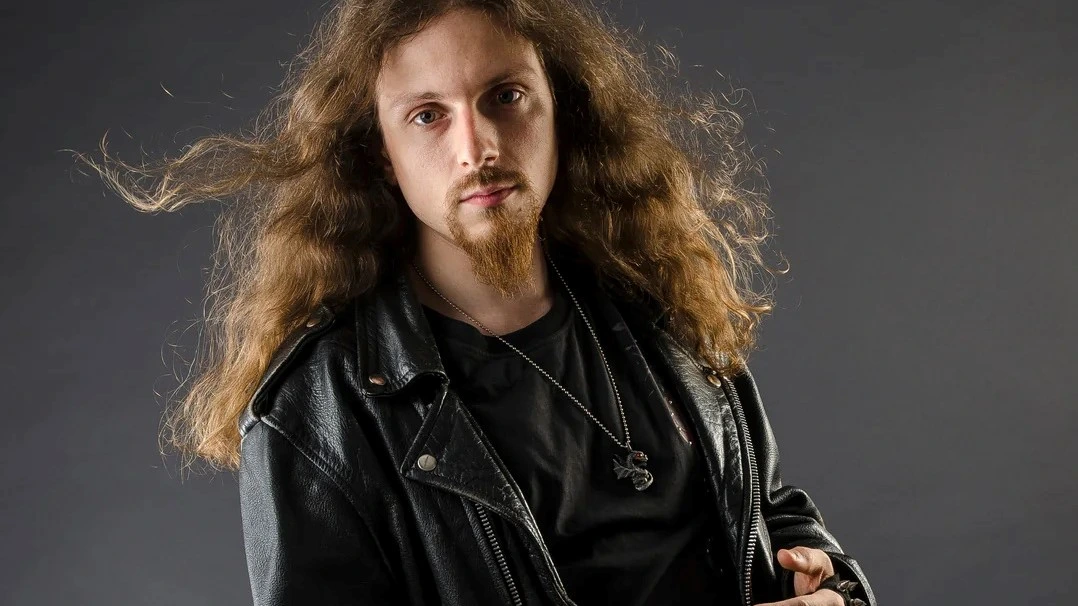 A man with long curly hair and a goatee wearing a black leather jacket against a grey backdrop, exuding a classic rocker vibe.