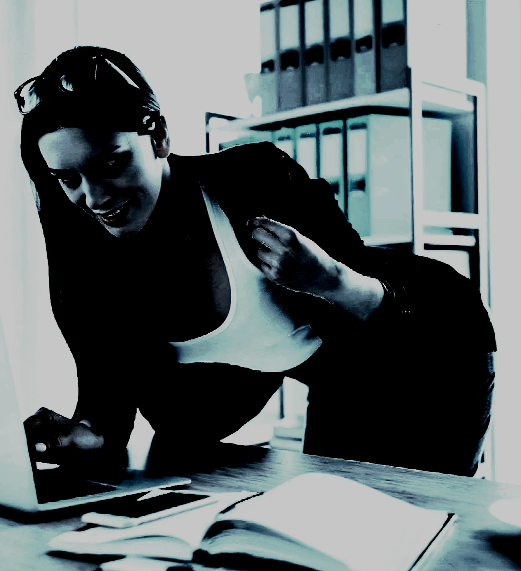 Woman leaning over a desk, smiling with a confident look, dressed in business attire in a softly lit office setting.
