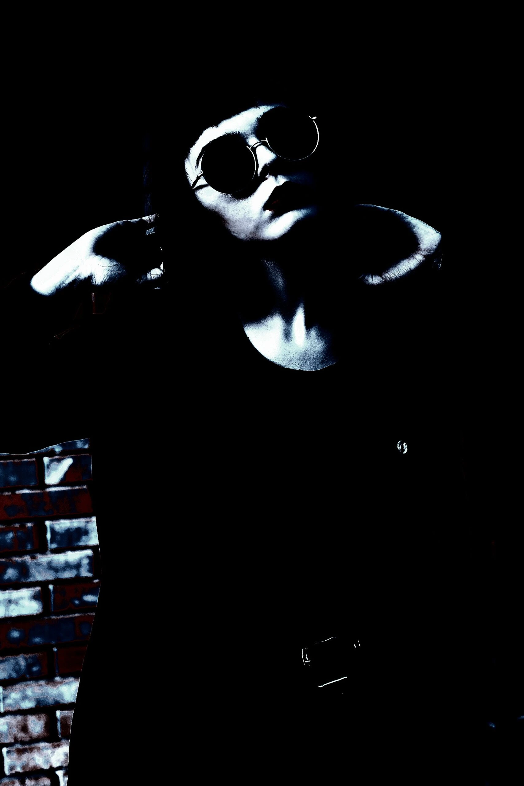 Individual wearing round sunglasses and dark clothing, standing against a dimly lit wall with an intense expression.
