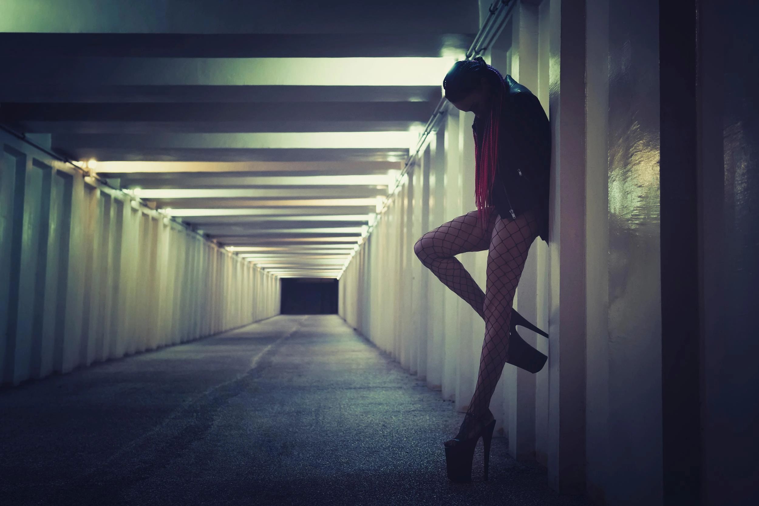 Woman in fishnet stockings and high heels leaning against a wall in a dimly lit urban tunnel, lost in thought.