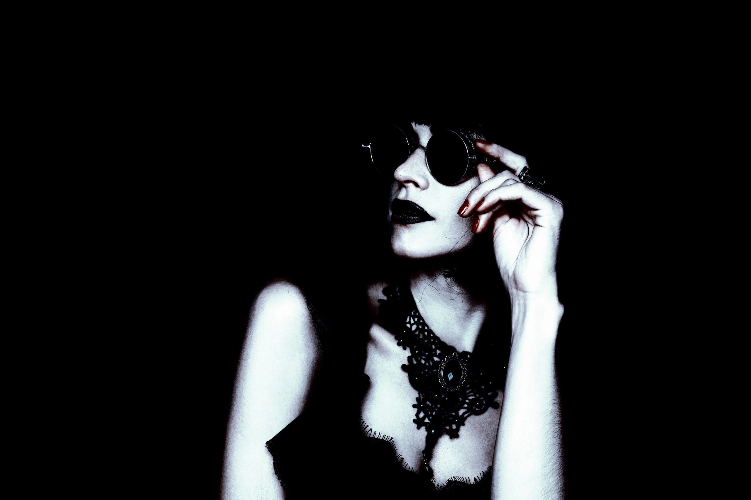 Woman in dark sunglasses and lace choker, gazing to the side in moody lighting with a dramatic, gothic style.