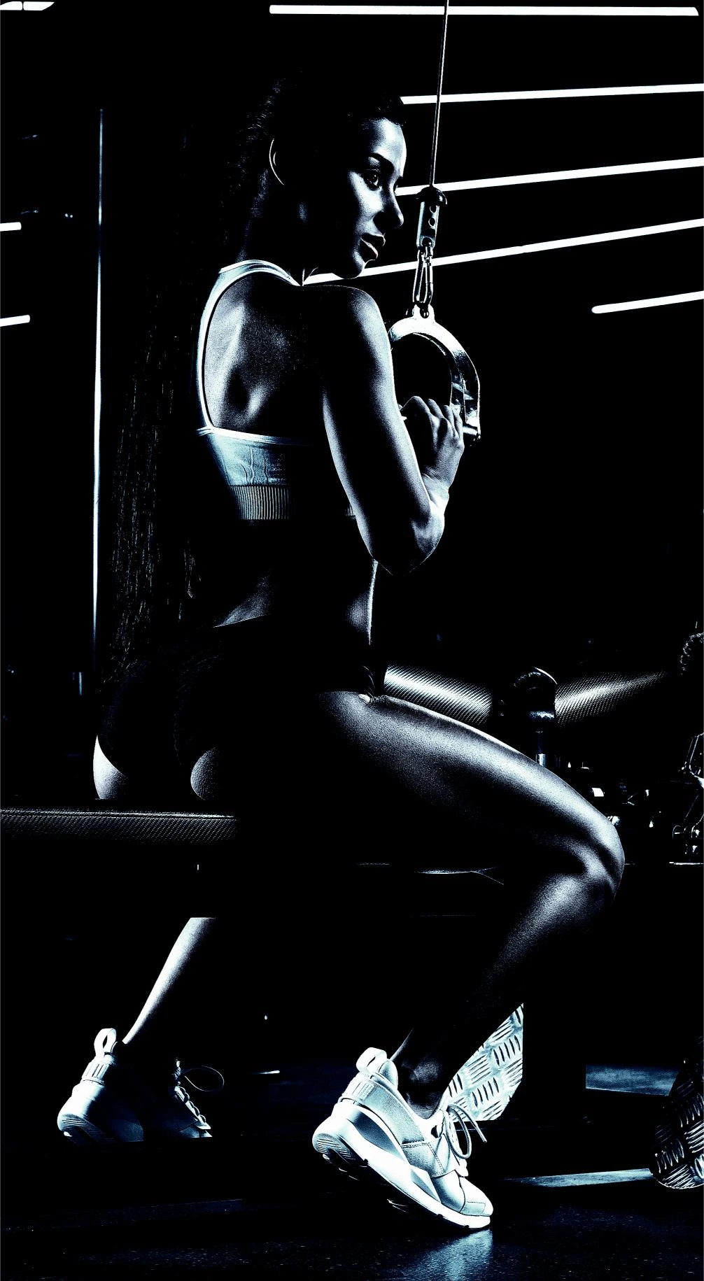 A woman seated at a gym machine in low light, gripping a handle with her back slightly turned, showcasing her toned back and arms.