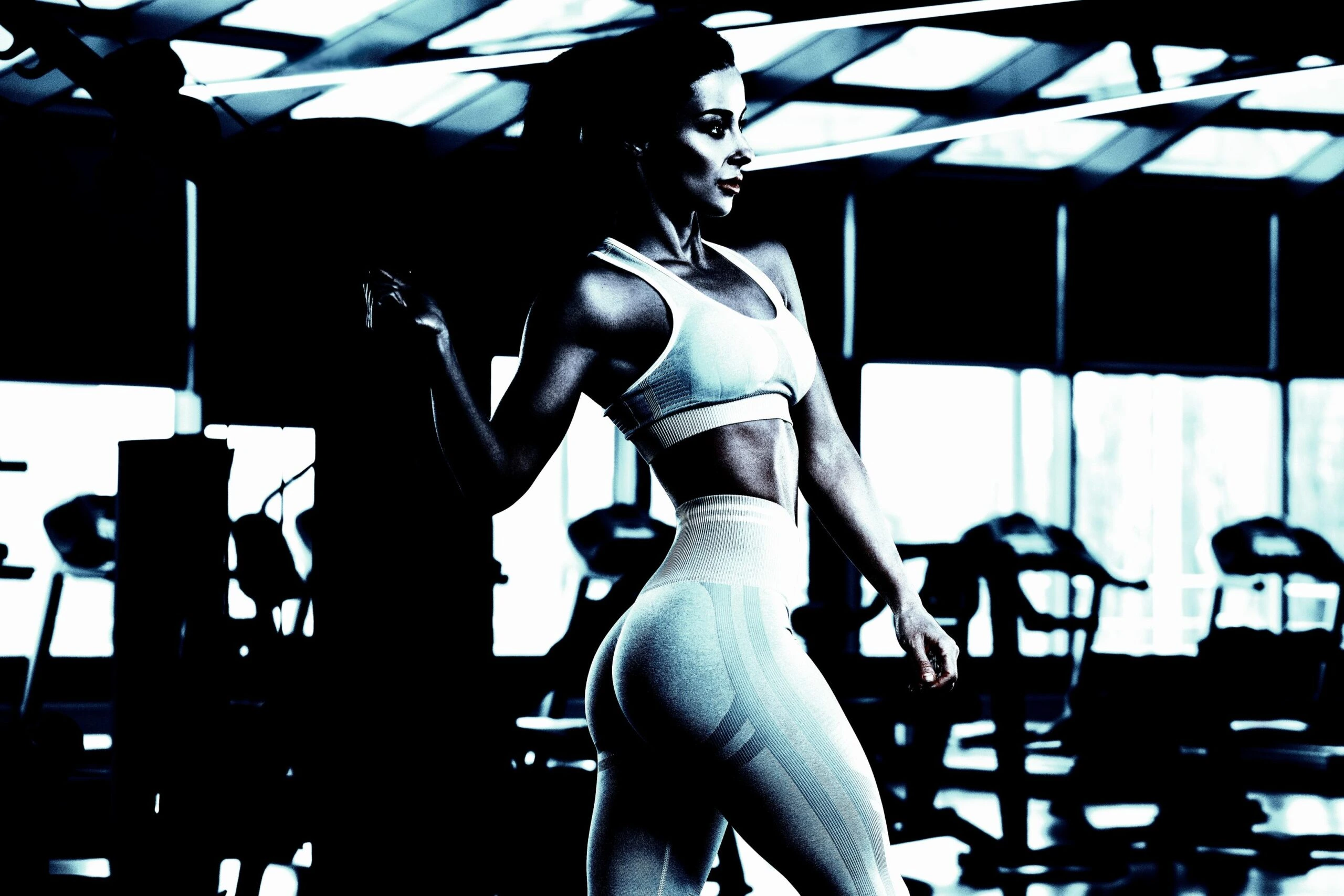 A fit woman standing in a gym, wearing a sports bra and leggings, with a strong, defined body under moody lighting.