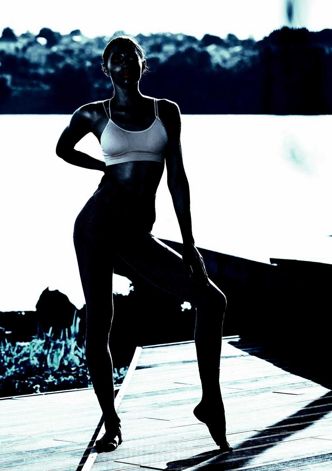 Silhouette of a woman in workout attire standing outdoors by the water, showcasing her toned body against a scenic background.