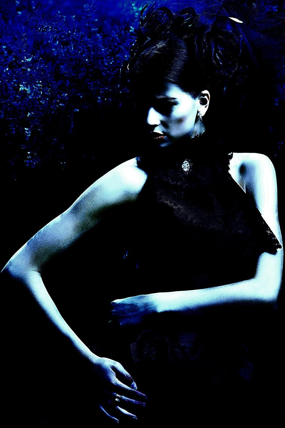 Woman sitting on a modern red chair in a black, textured gothic gown, with a brooding expression in an industrial setting.