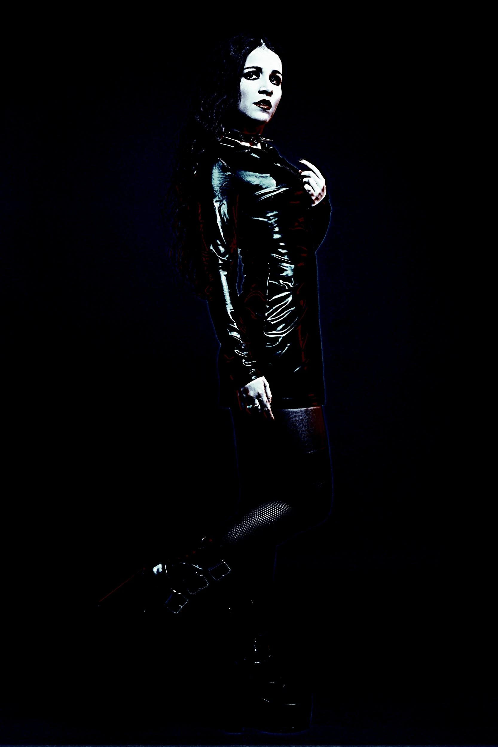 Woman in a black, glossy latex dress with a dark, intense gaze and long black hair, exuding gothic elegance against a dark background.