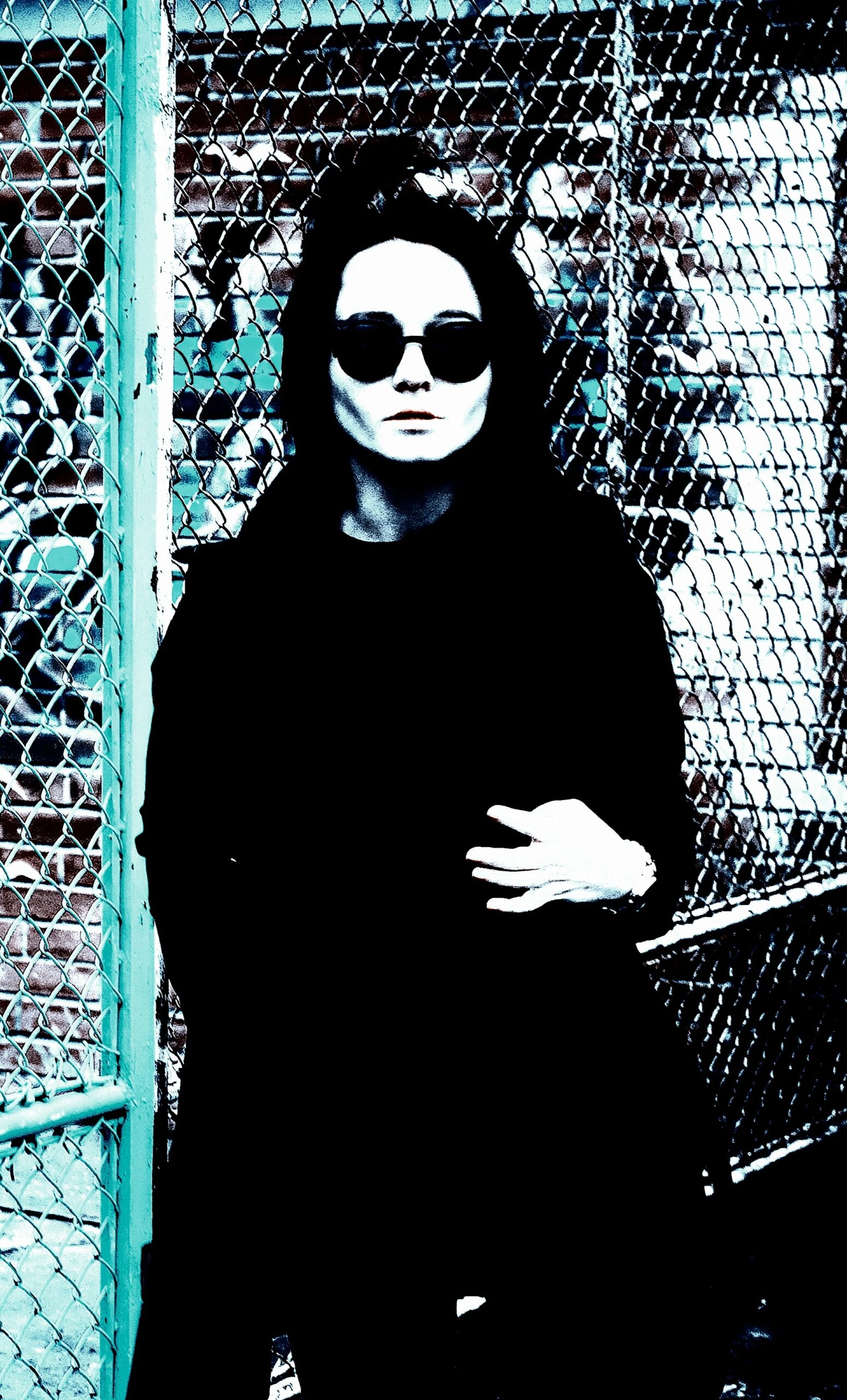 Person in black coat and dark sunglasses standing against a chain-link fence, capturing an urban, edgy look.