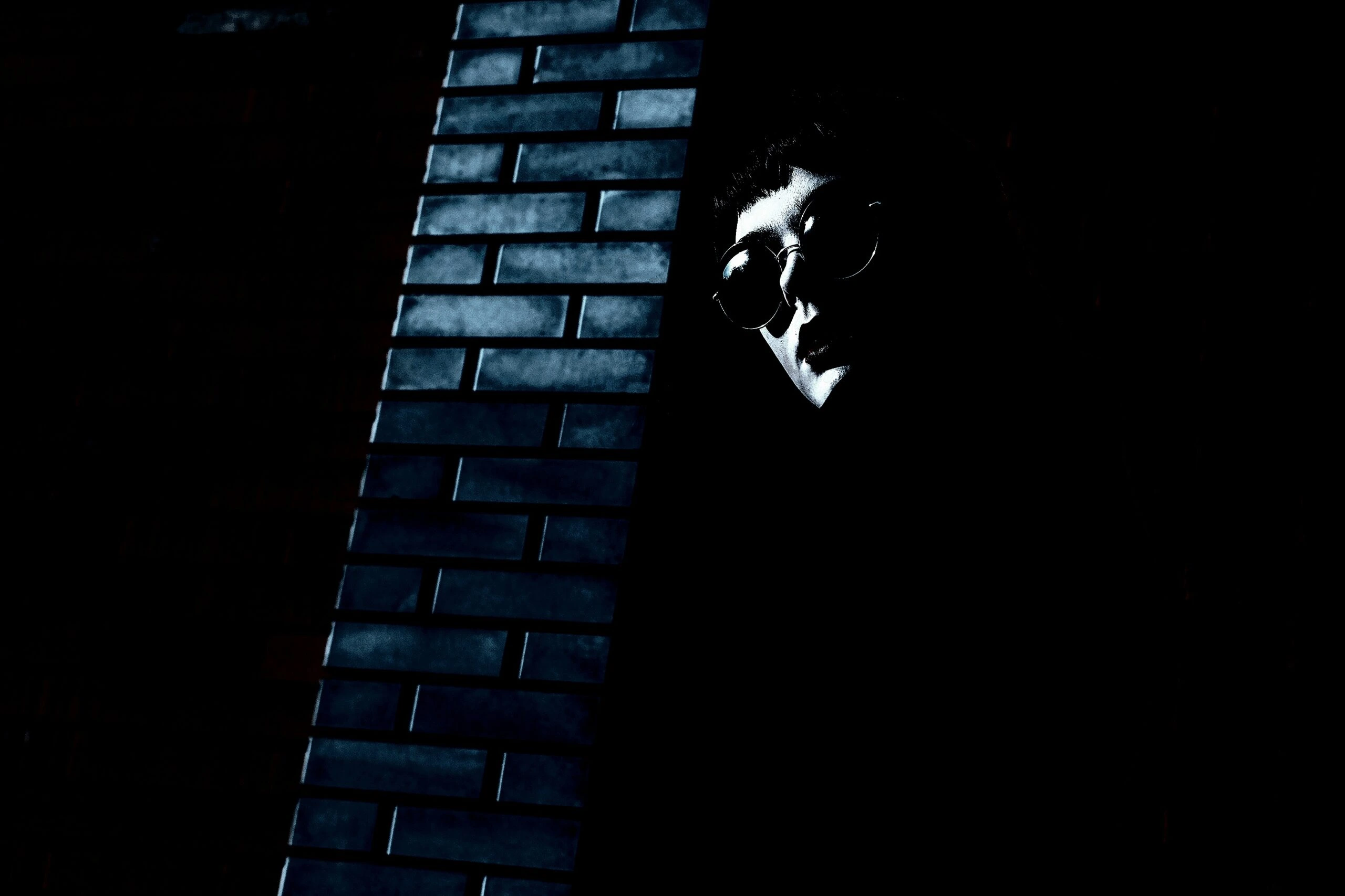 Person in sunglasses partially illuminated against a dark brick wall, exuding a mysterious rock aesthetic.