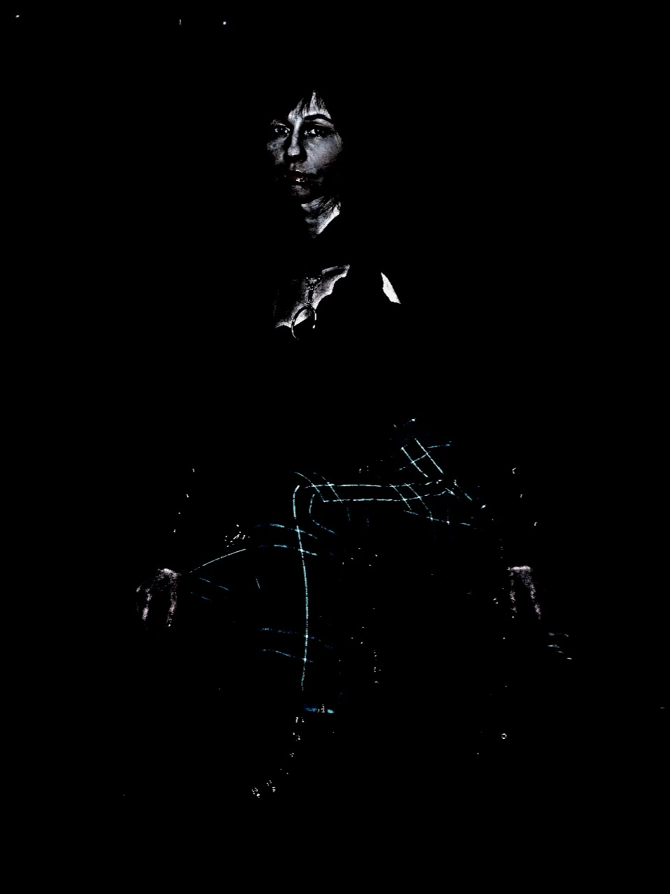 A figure emerges from darkness, face illuminated, dressed in dark clothing with vivid blue streaks, creating an enigmatic presence.