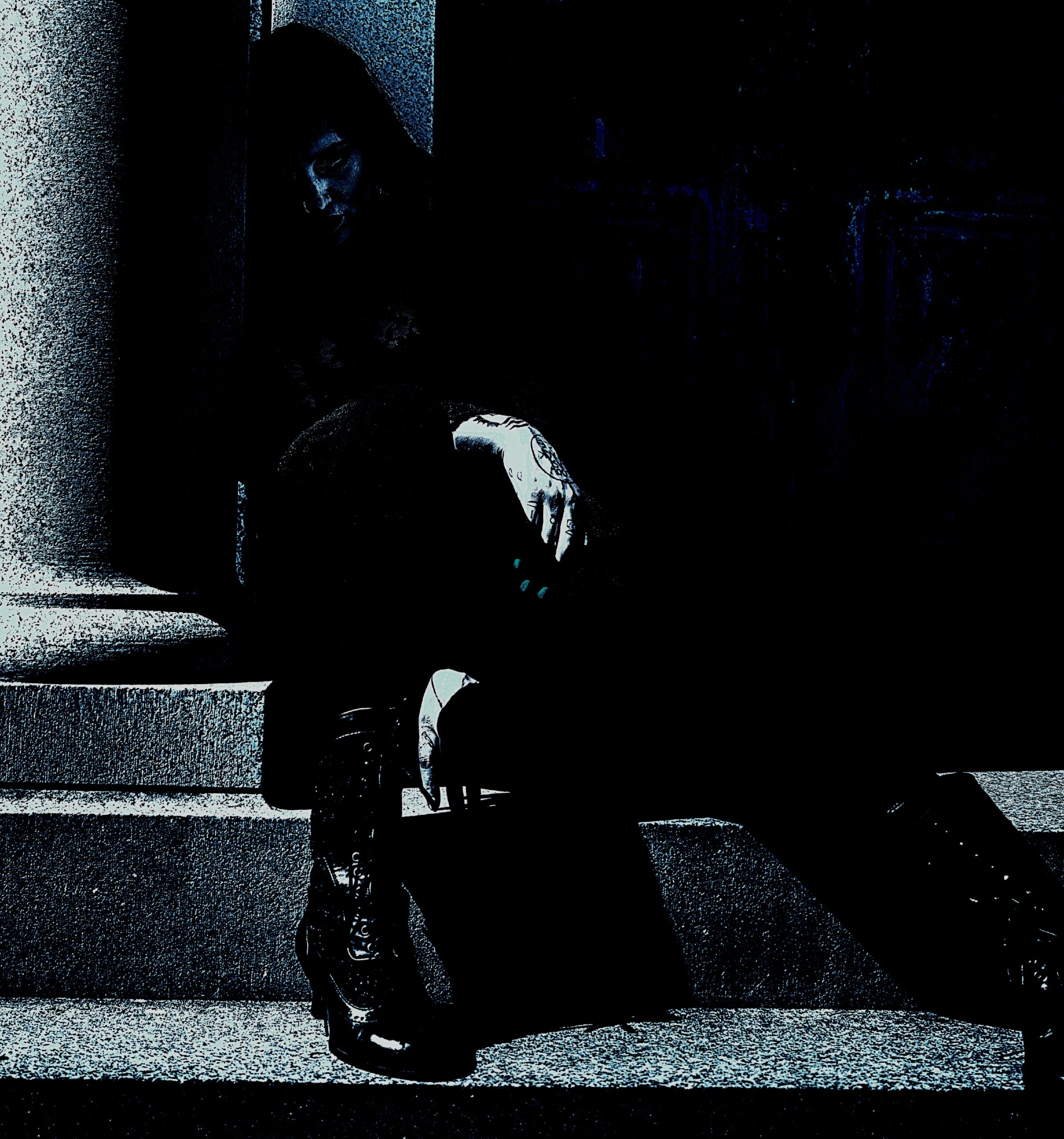 A solitary figure sits on stone steps, shrouded in shadows, wearing dark pants with bold lacing on the sides, embodying a gothic aesthetic with chunky, high-heeled boots.
