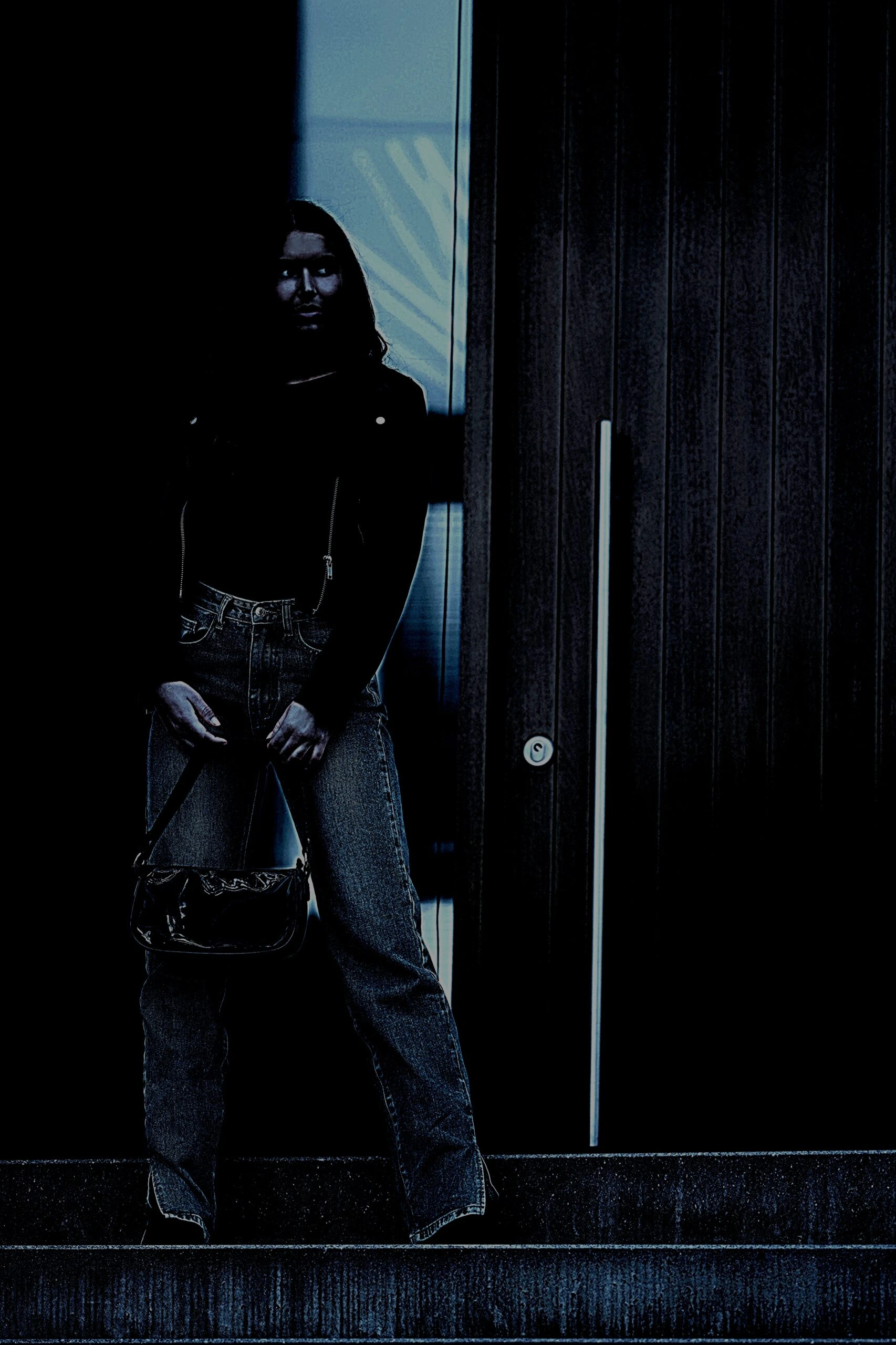 A person stands in a dimly lit doorway, sporting classic straight-legged denim jeans and a black top, complemented by a dark, shiny shoulder bag, merging casual with goth influences.