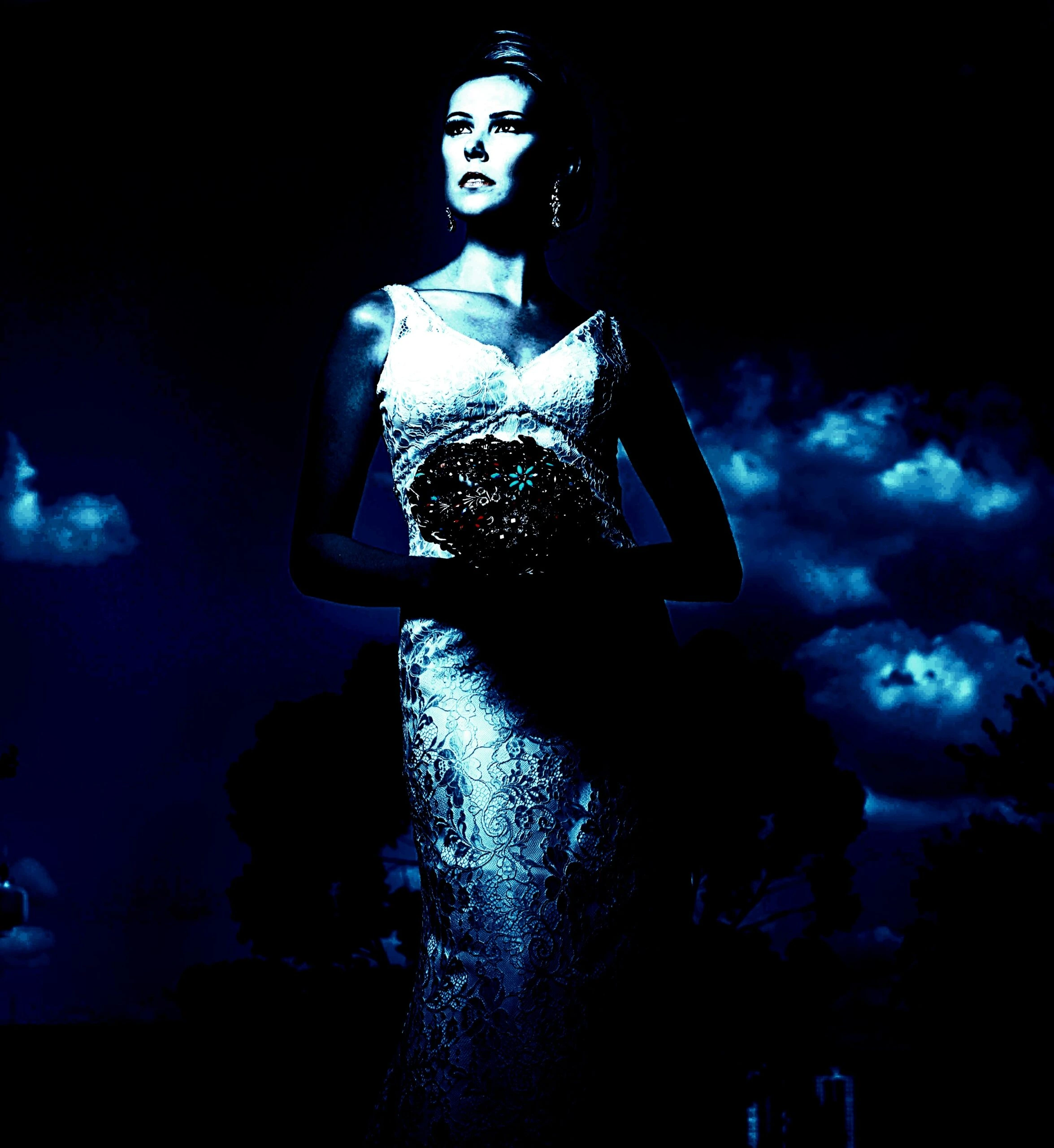 A gothic bride in a blue-toned photograph stands against a night sky, the dress detailed with lace, embodying a blend of elegance and mystery.