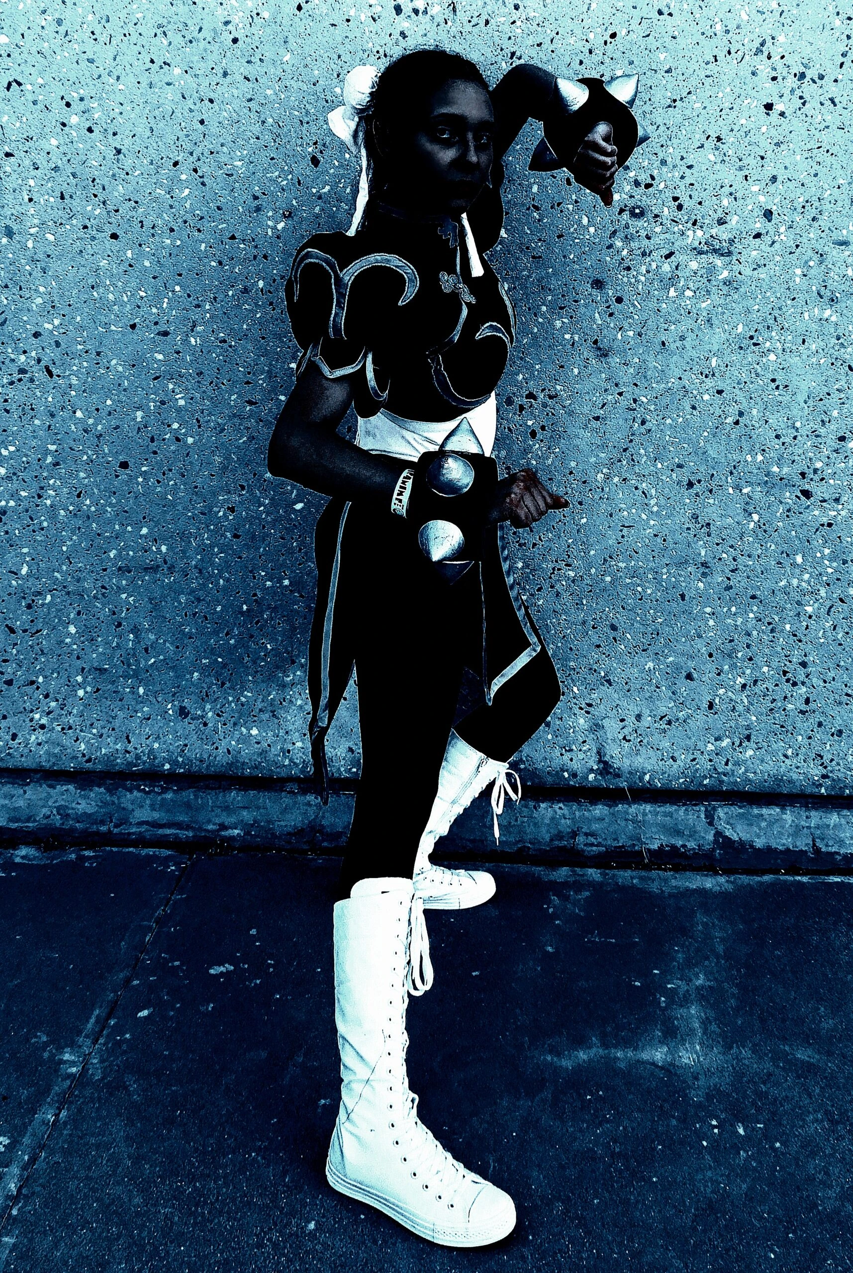 Cosplayer in a black and white costume posing confidently on a textured pavement, with a dramatic blue color cast.