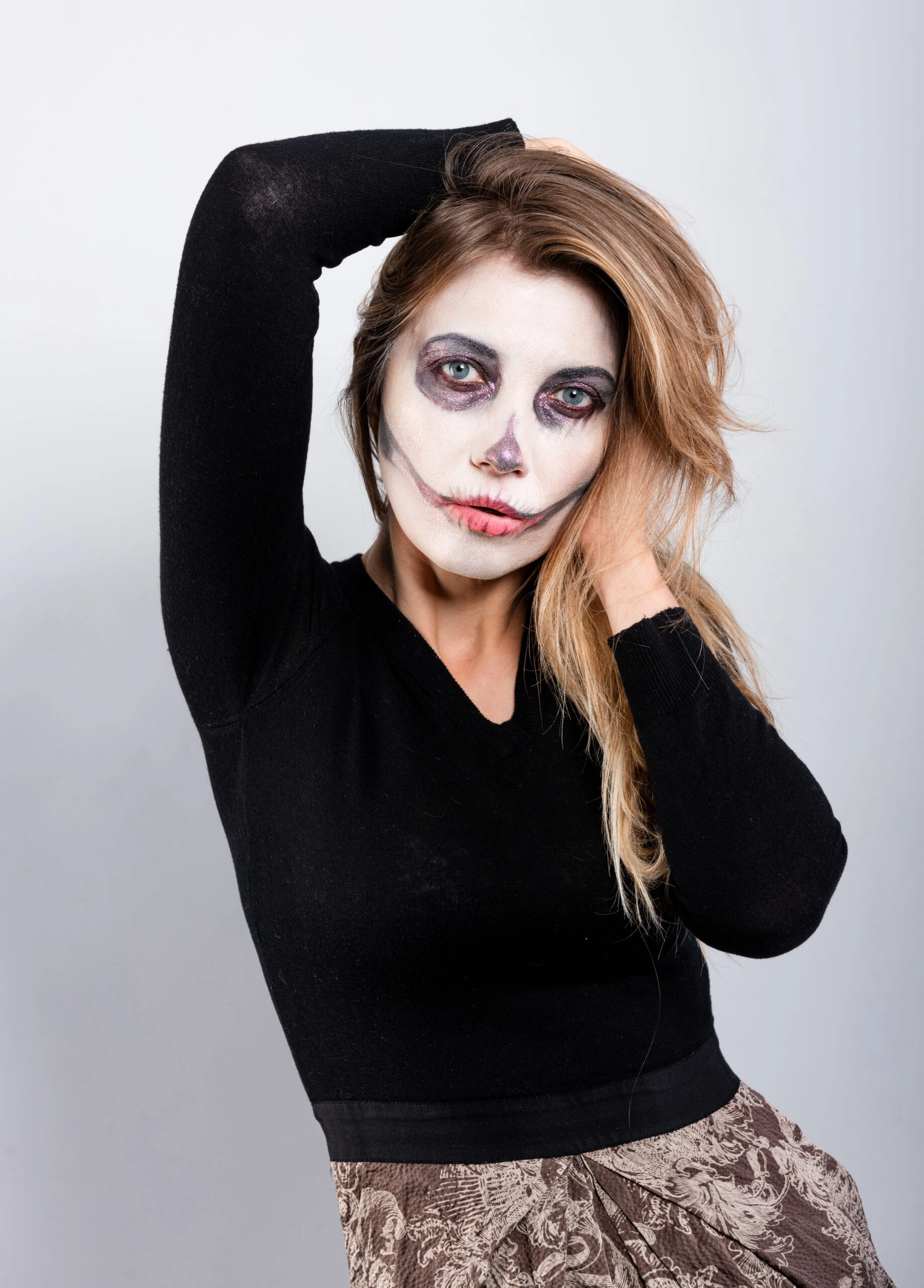 A woman with gothic skull makeup in a black sweater, posing with her hand behind her head.