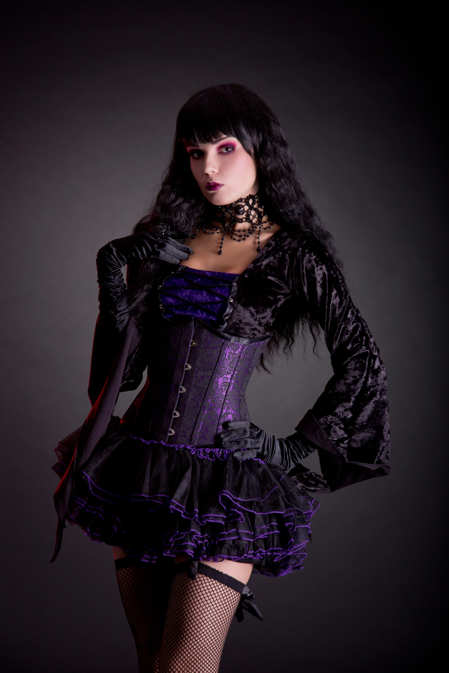 A woman with long black hair wearing a goth-inspired outfit with a purple and black corset, black velvet jacket, ruffled skirt, and fishnet stockings, poised in a dramatic stance against a dark background.