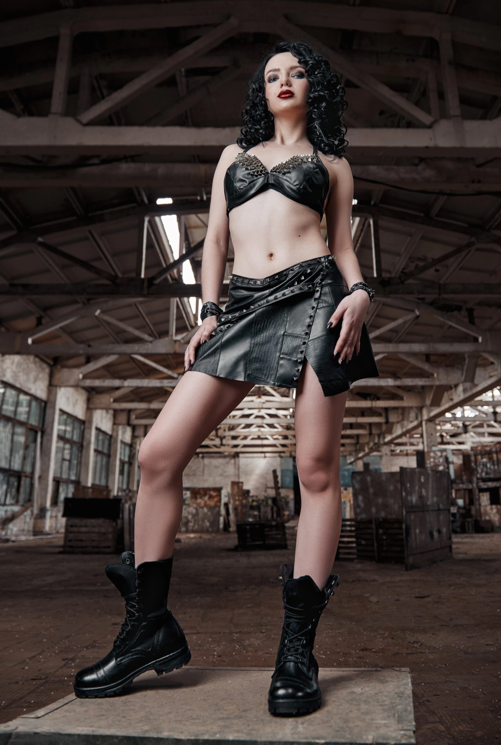 Full-body shot of a woman with black curly hair, in a black leather outfit complete with studded details and heavy black boots, standing with an assertive posture in an abandoned industrial setting.