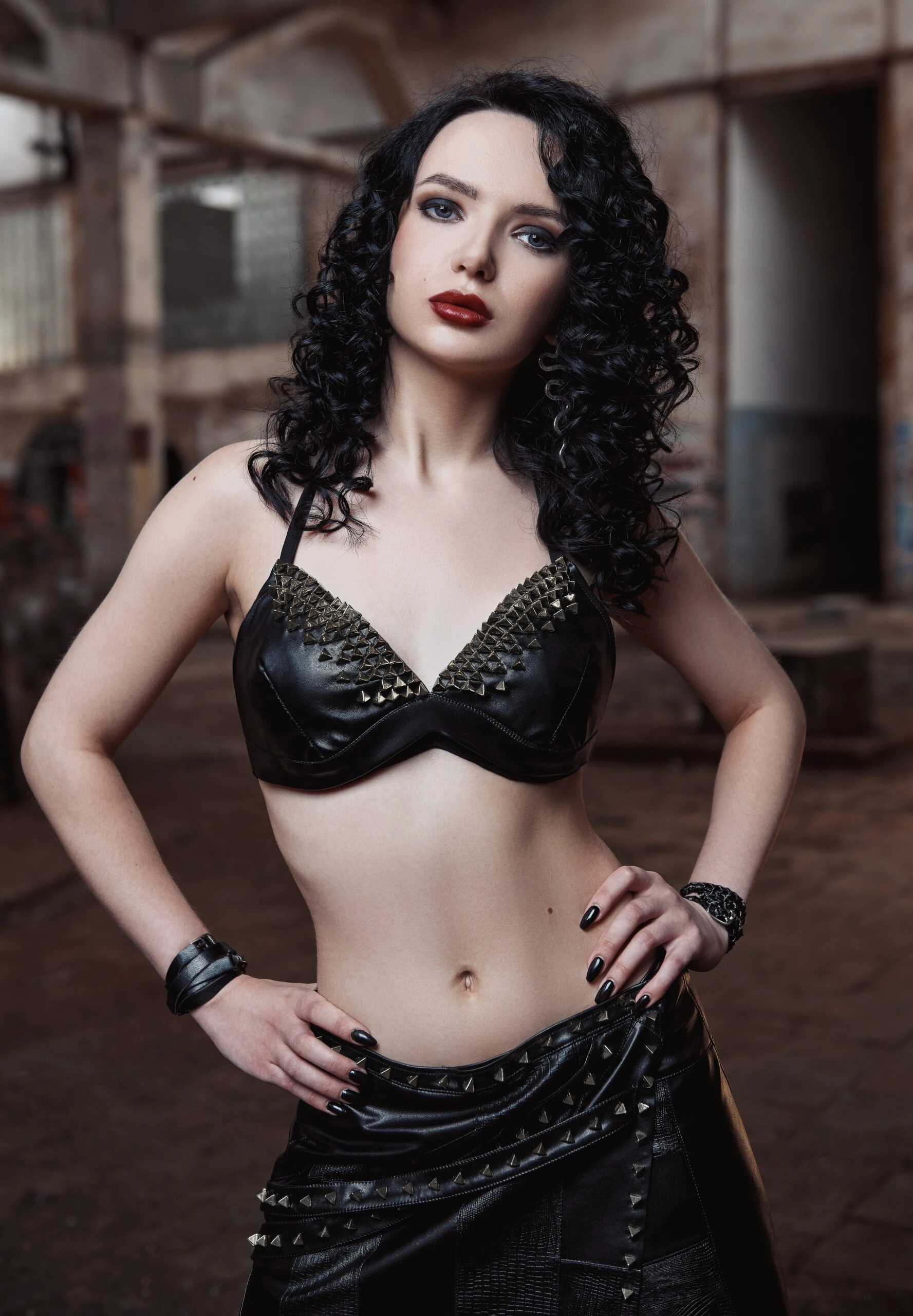 A confident woman with porcelain skin and jet-black curly hair, dressed in a studded black leather bralette and a layered skirt adorned with metallic spikes, stands in an industrial setting with a resolute stance.