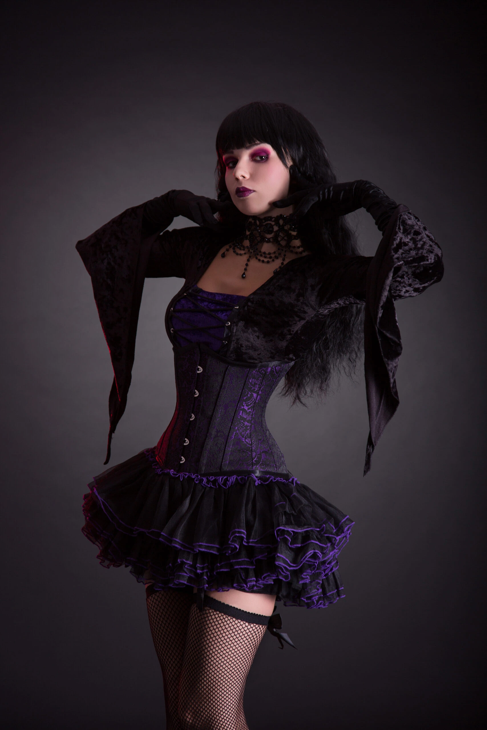 Goth woman striking a confident pose with one hand on her waist, her black velvet jacket's sleeve draping elegantly, complementing her corset and skirt ensemble.