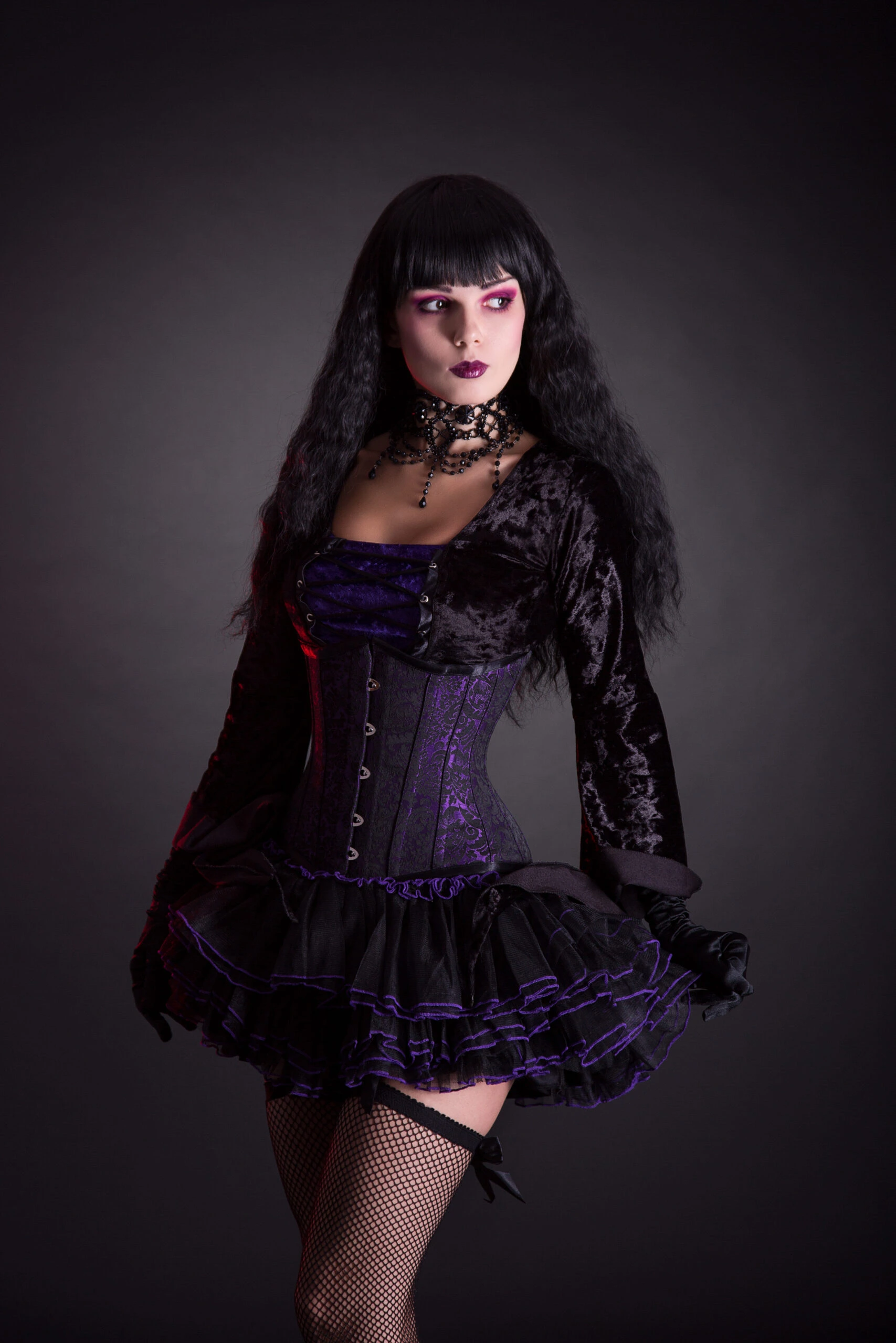 Woman in a gothic costume holding a black top hat in one hand, standing confidently with her other hand on her hip, showcasing her vibrant makeup and layered choker.