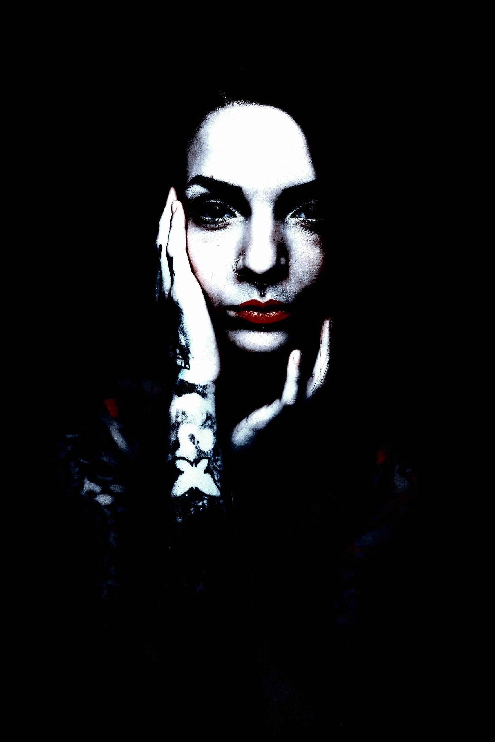 A high-contrast portrait of a person with a serene expression, adorned with a skull tattoo on the forearm and an occult-style necklace, set against a dark background.