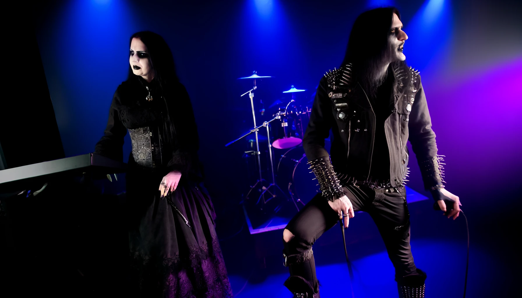 Eerie stage scene with a gothic duo, woman in black lace and man in spiky attire, playing synths in a foggy, dimly lit setting. Ashbury Heights - SmALLer.