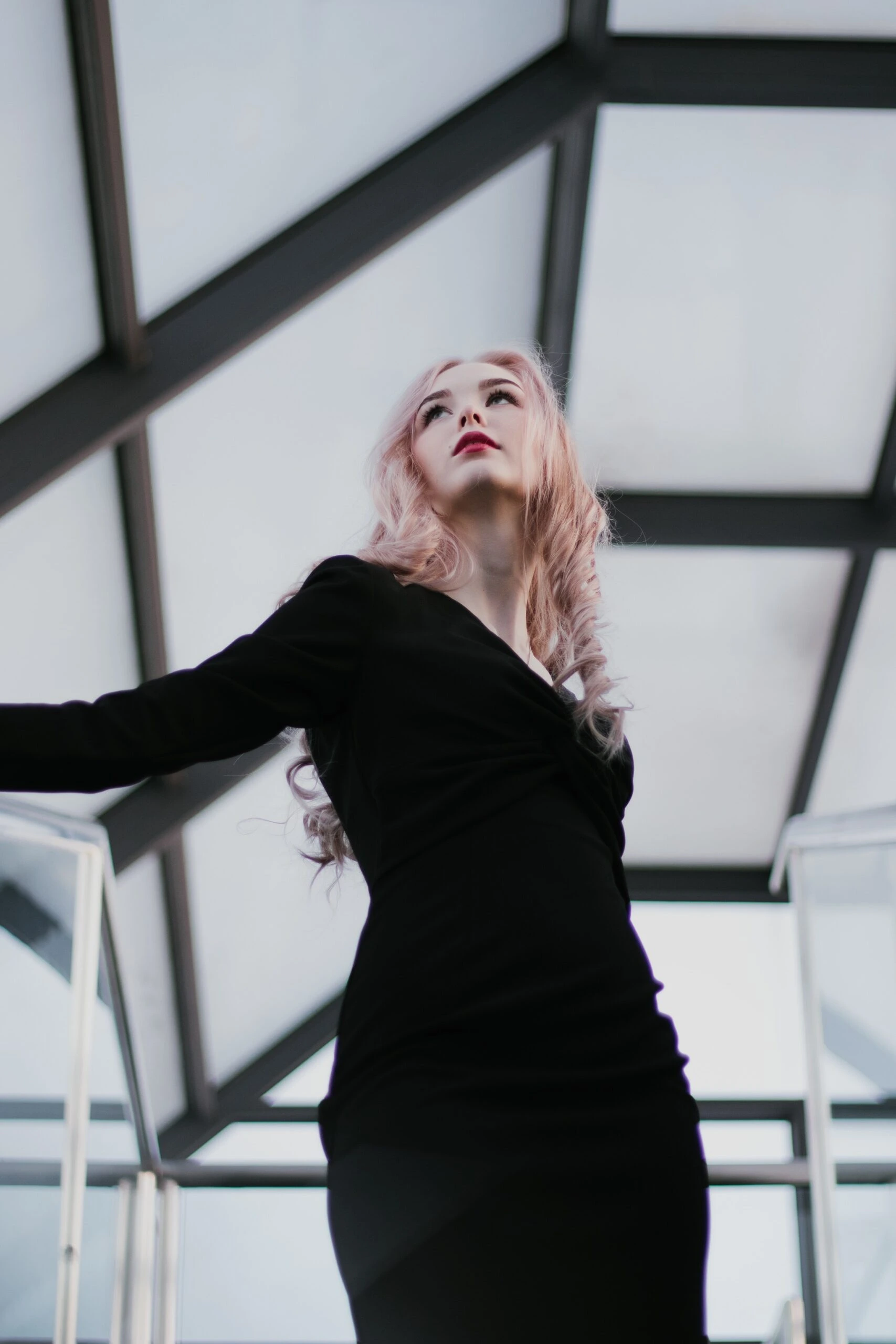 Young woman in a sleek black dress looking upwards in a modern glass building.