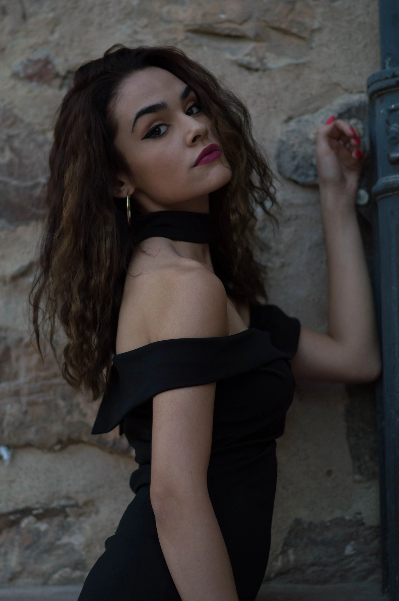 Woman in a chic off-shoulder black dress posing against an old stone wall.