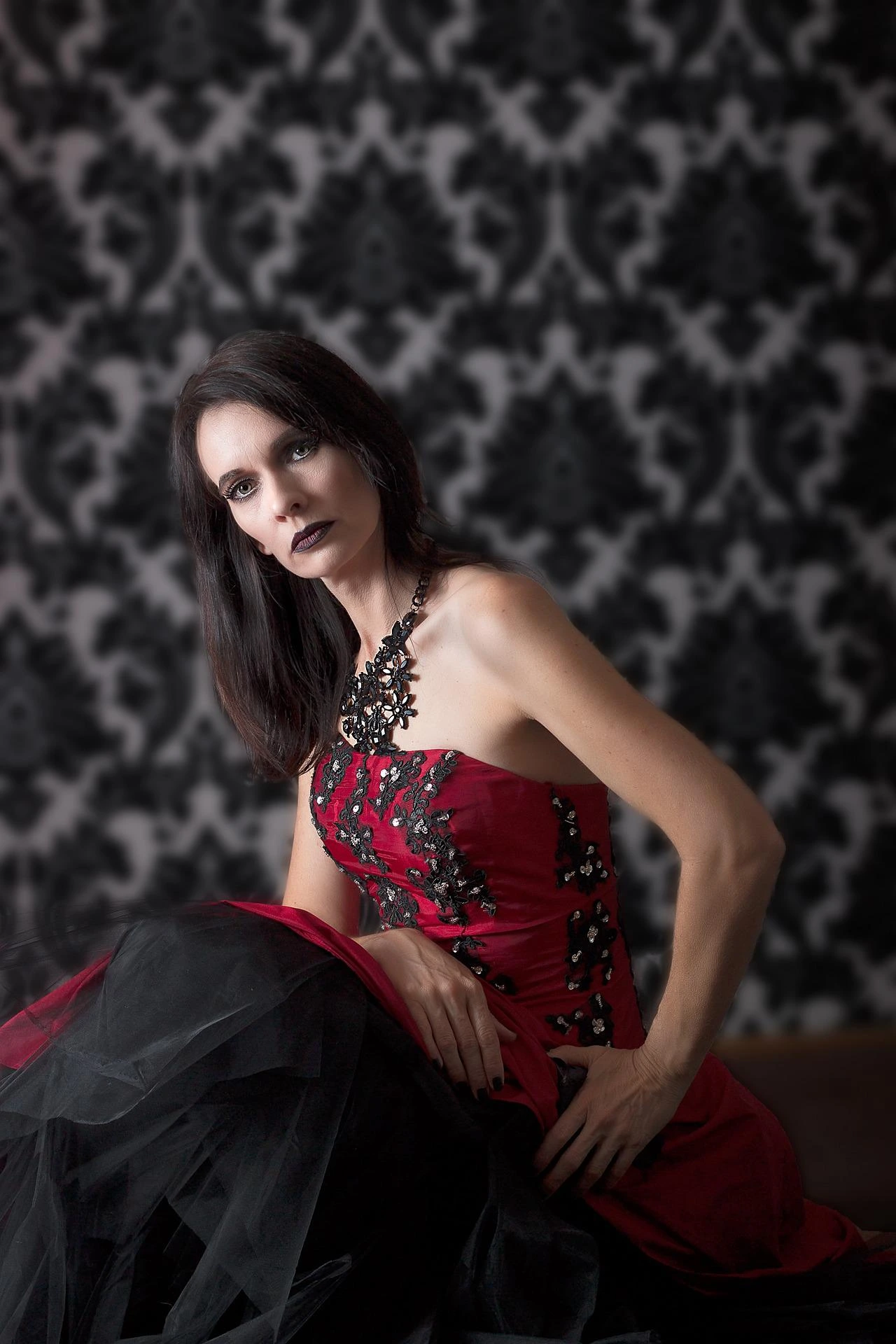 Gothic woman in a red and black dress with elaborate embroidery sitting against a patterned backdrop.