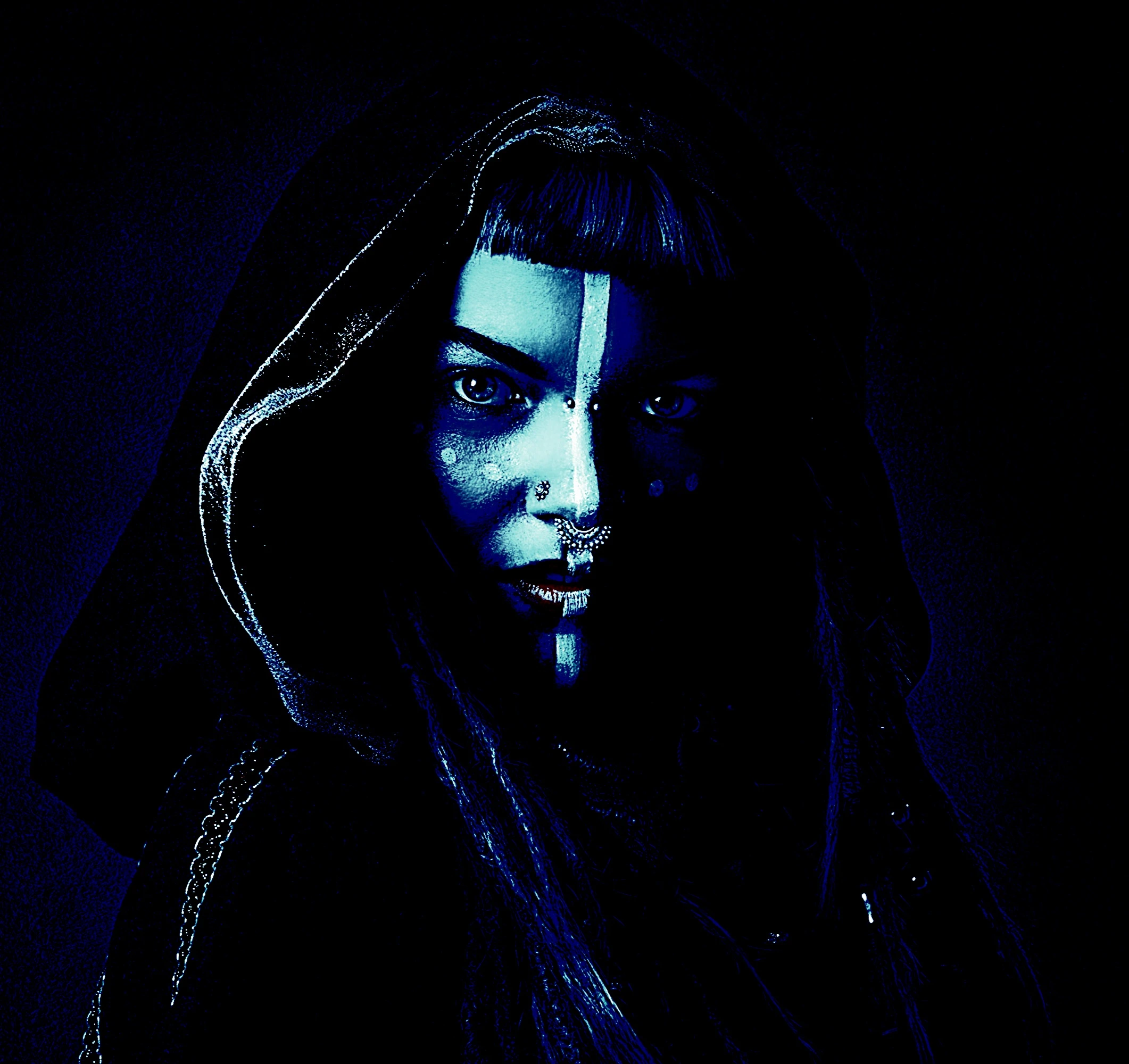 Intense gothic makeup with blue hues on a woman wearing a hood, featuring facial piercings and mystical face paint.