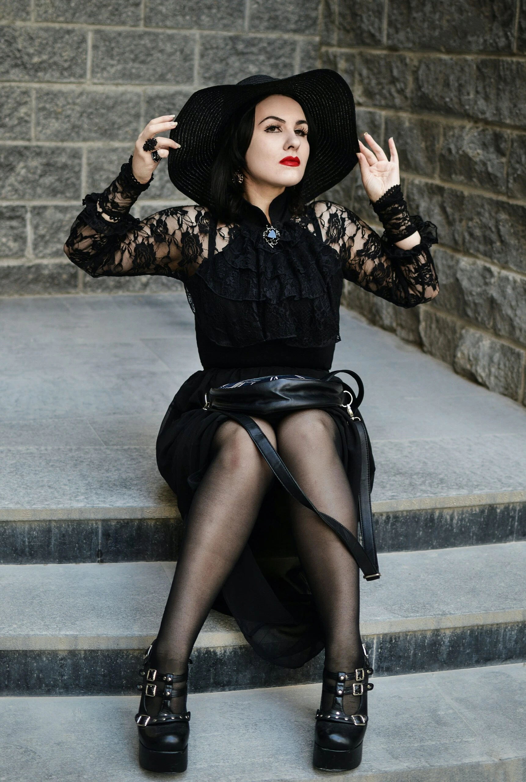 Gothic fashion model in a black lace dress and wide-brimmed hat seated on steps.
