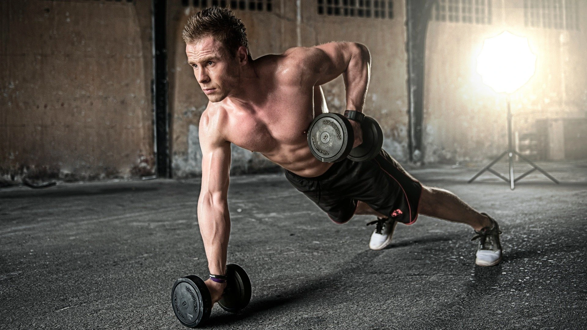 Man performing one-arm push-up with dumbbells in gritty workout space.