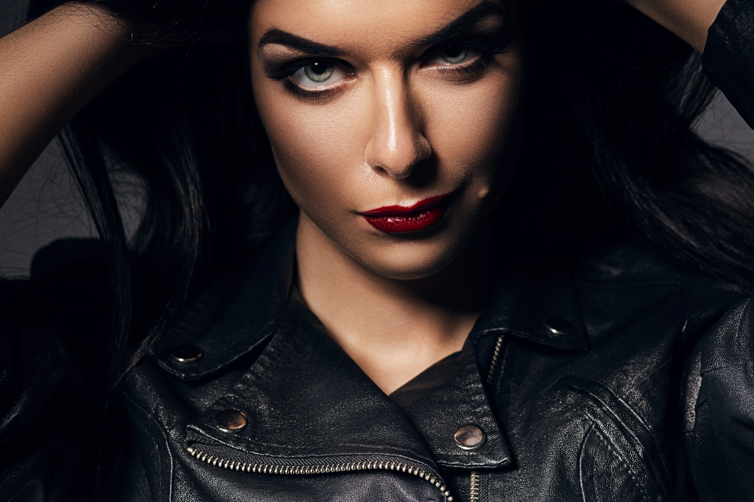A close-up of a woman with dark hair and striking makeup, featuring smokey eyes and red lipstick, wearing a leather jacket.