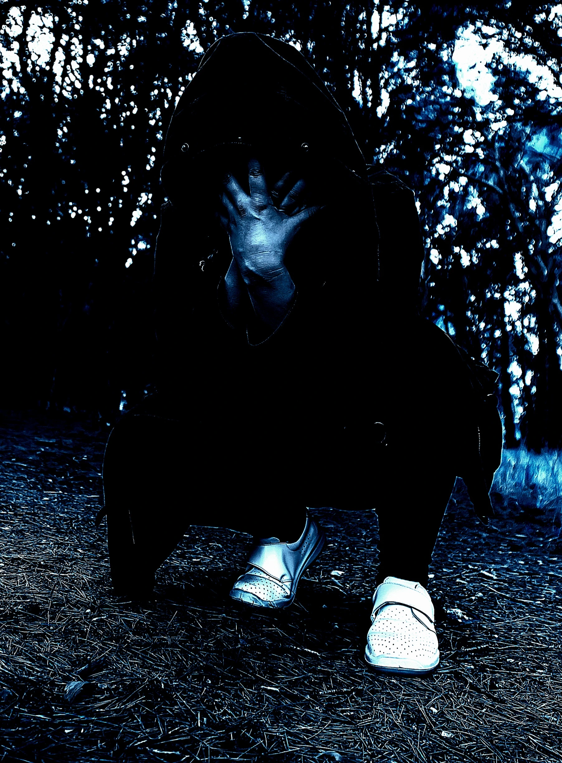 Shadowy figure in a gothic outfit hiding face with hands, set against a background of blurred forest foliage.