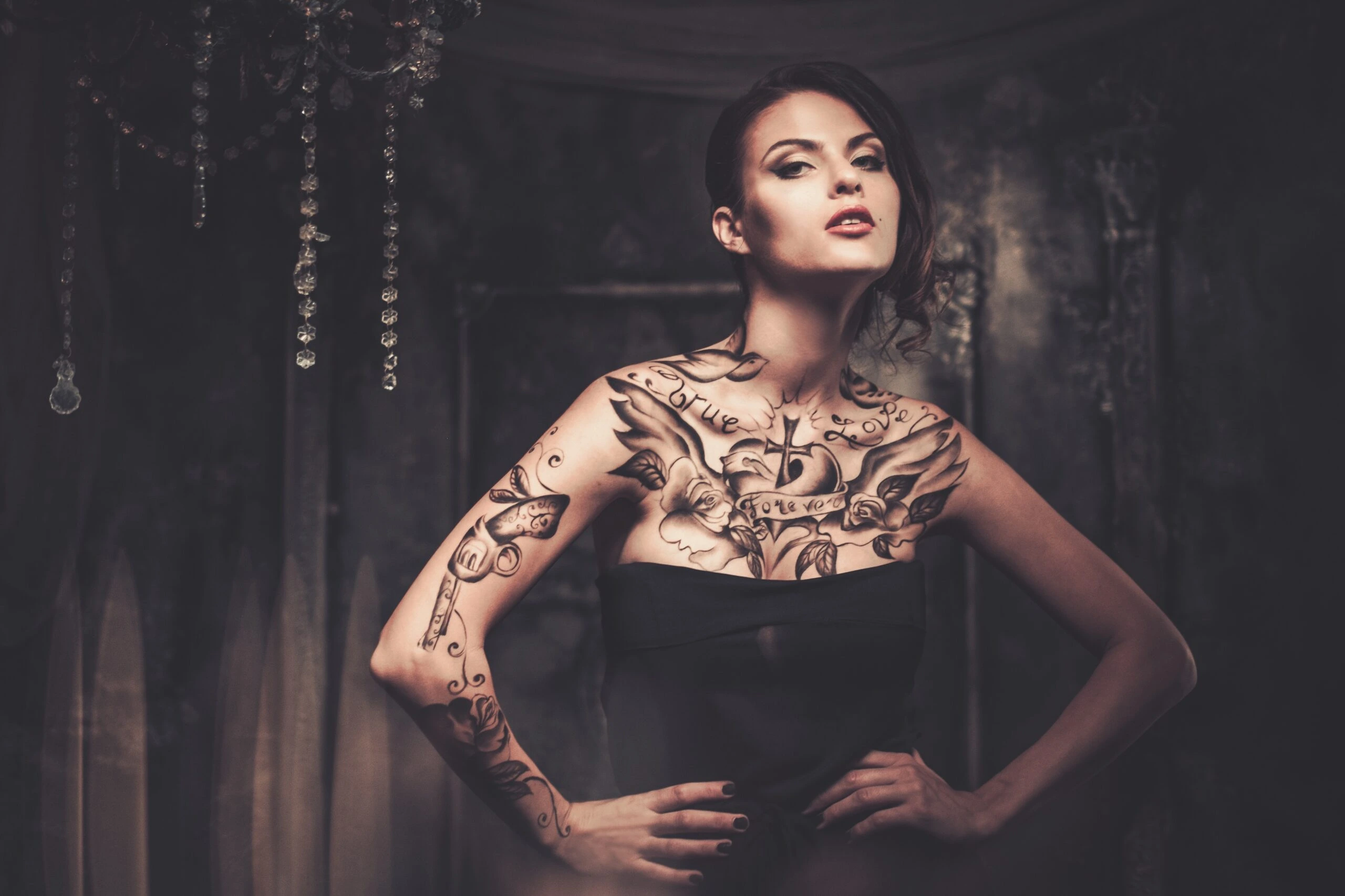 Elegant tattooed woman in a dark setting with intricate chest and arm tattoos, a deep look into Tattoos in the Metal Subculture.
