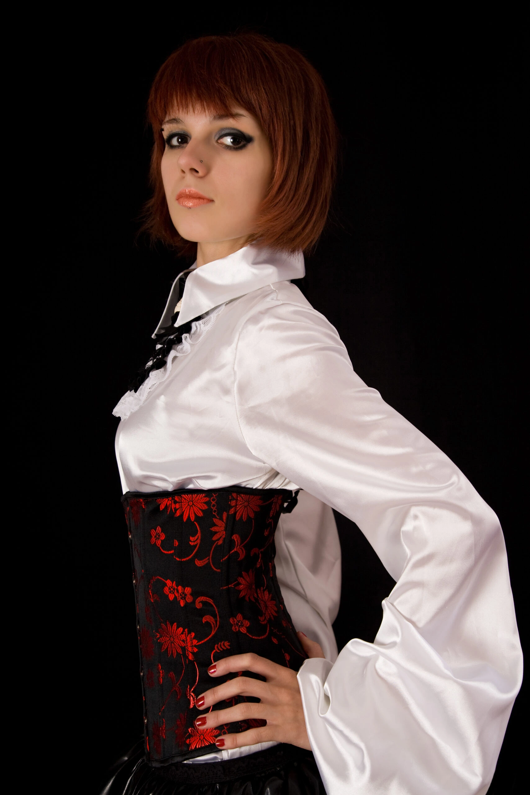 A woman with auburn hair in a short bob cut wearing a white blouse with a high collar and a black corset with red floral patterns.