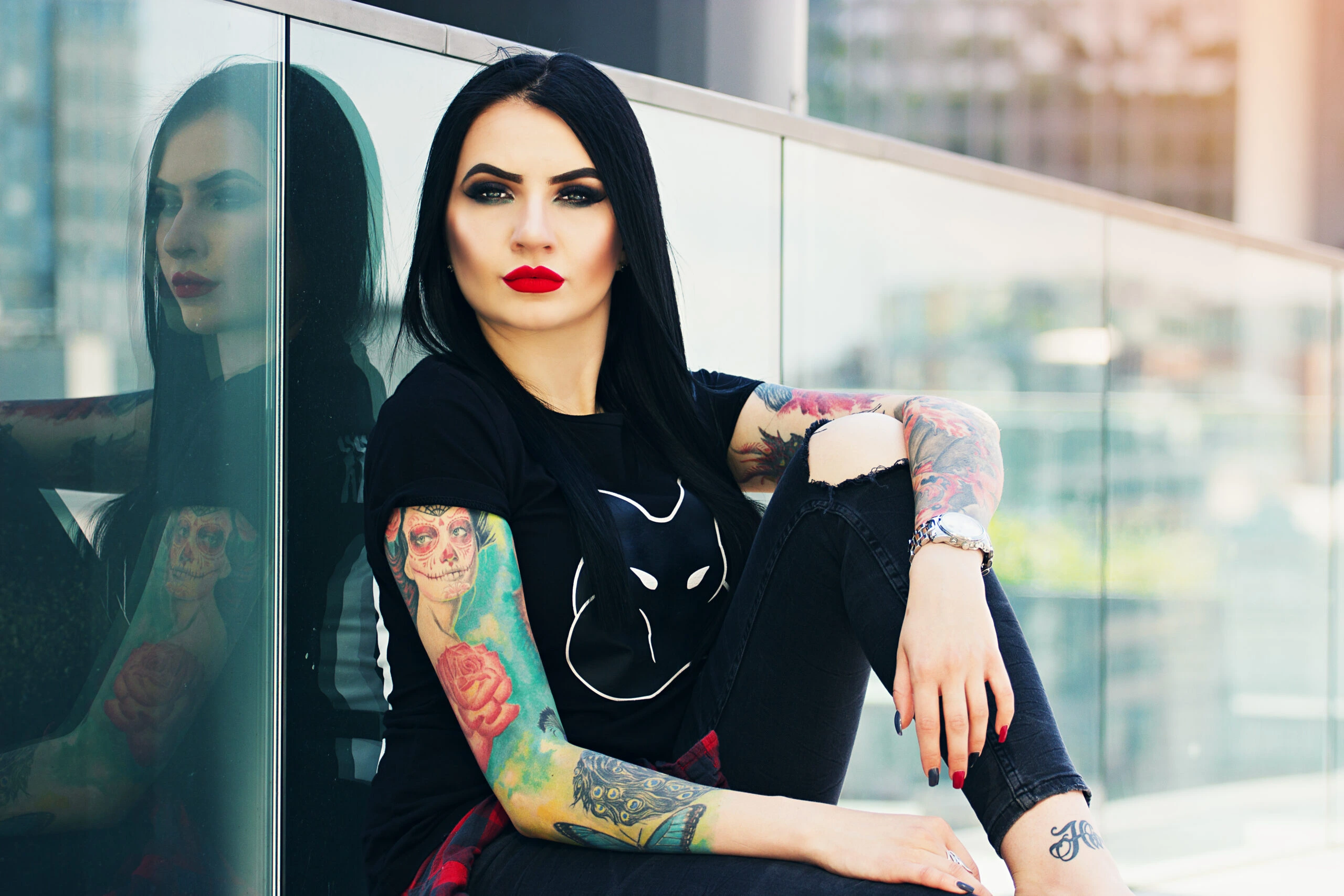A woman with black hair and tattoos, wearing a black t-shirt with a white alien print and distressed black jeans, sitting casually in front of a reflective glass building.