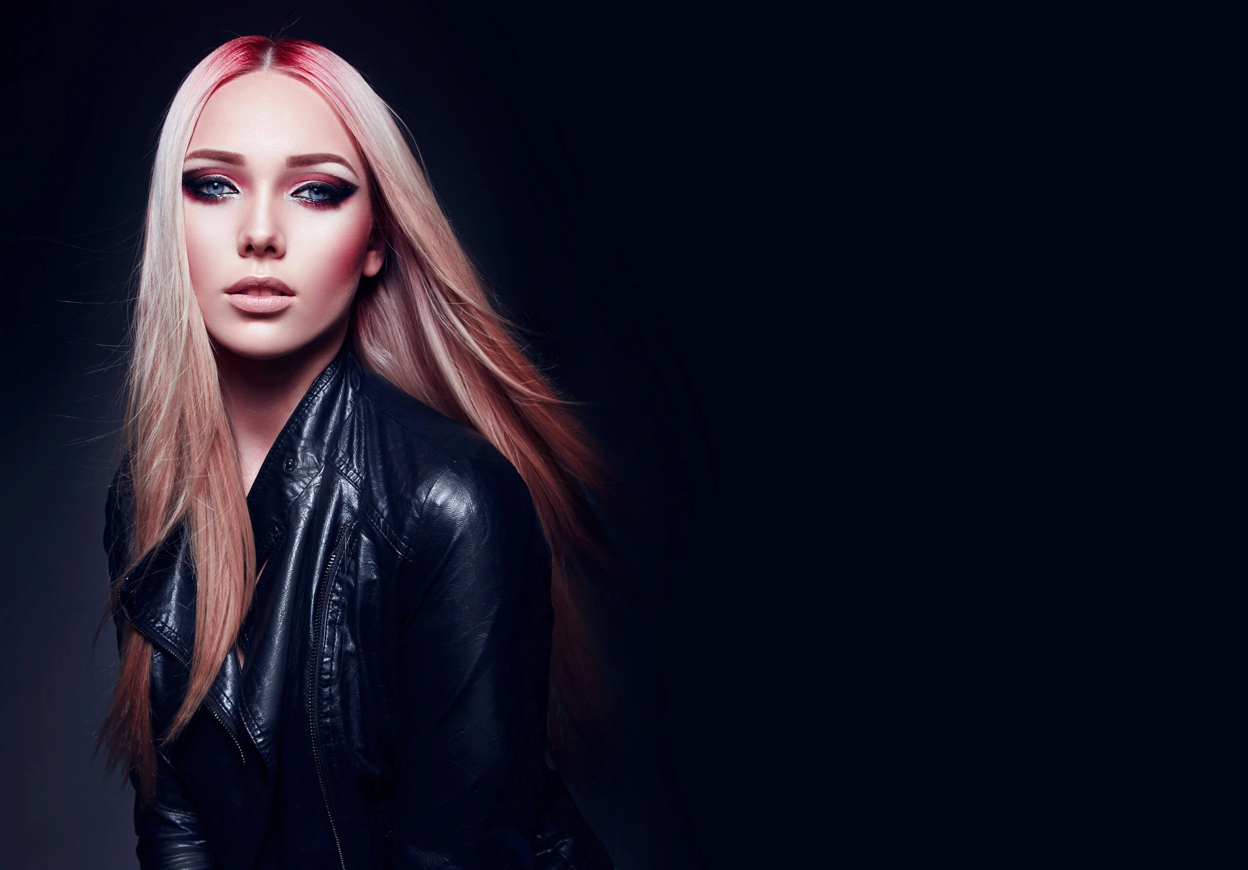 A woman with pink and blonde hair and dramatic eye makeup wearing a black leather jacket and looking pensively to the side.