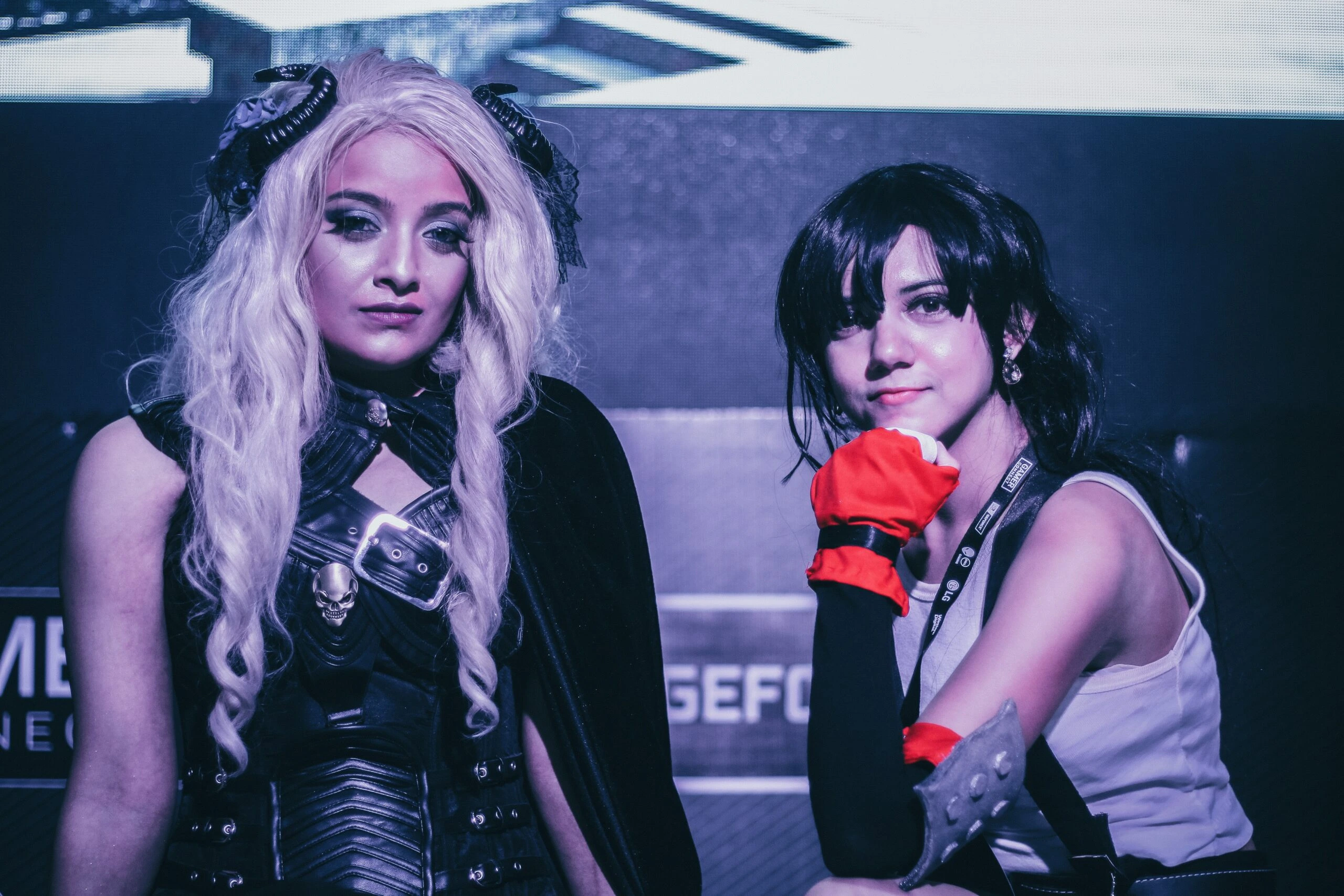 Two cosplayers posing at a gaming event, one in a dark fantasy outfit with long white hair and the other in a sporty costume with red gloves.