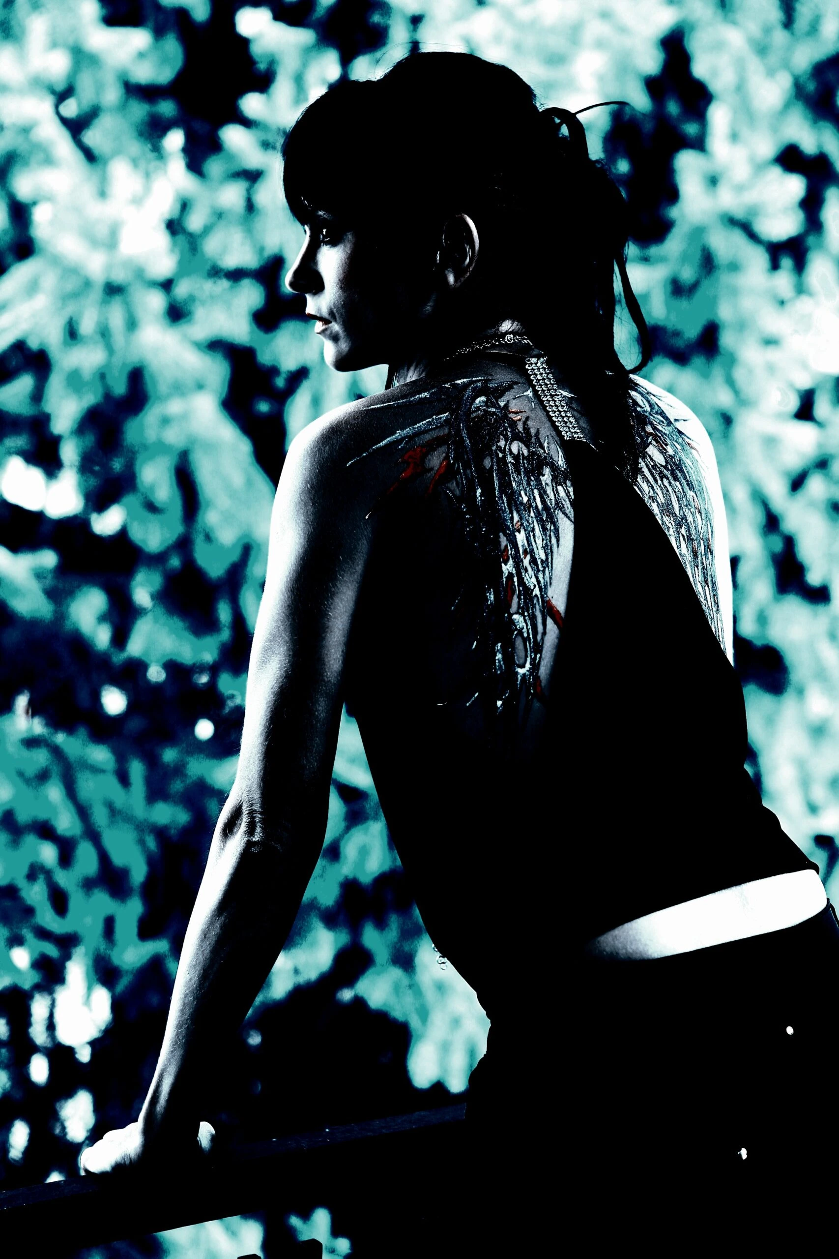 A gothic woman with dark hair and a tattoo of angel wings on her back, gazing off to the side against a blue-toned leafy background.
