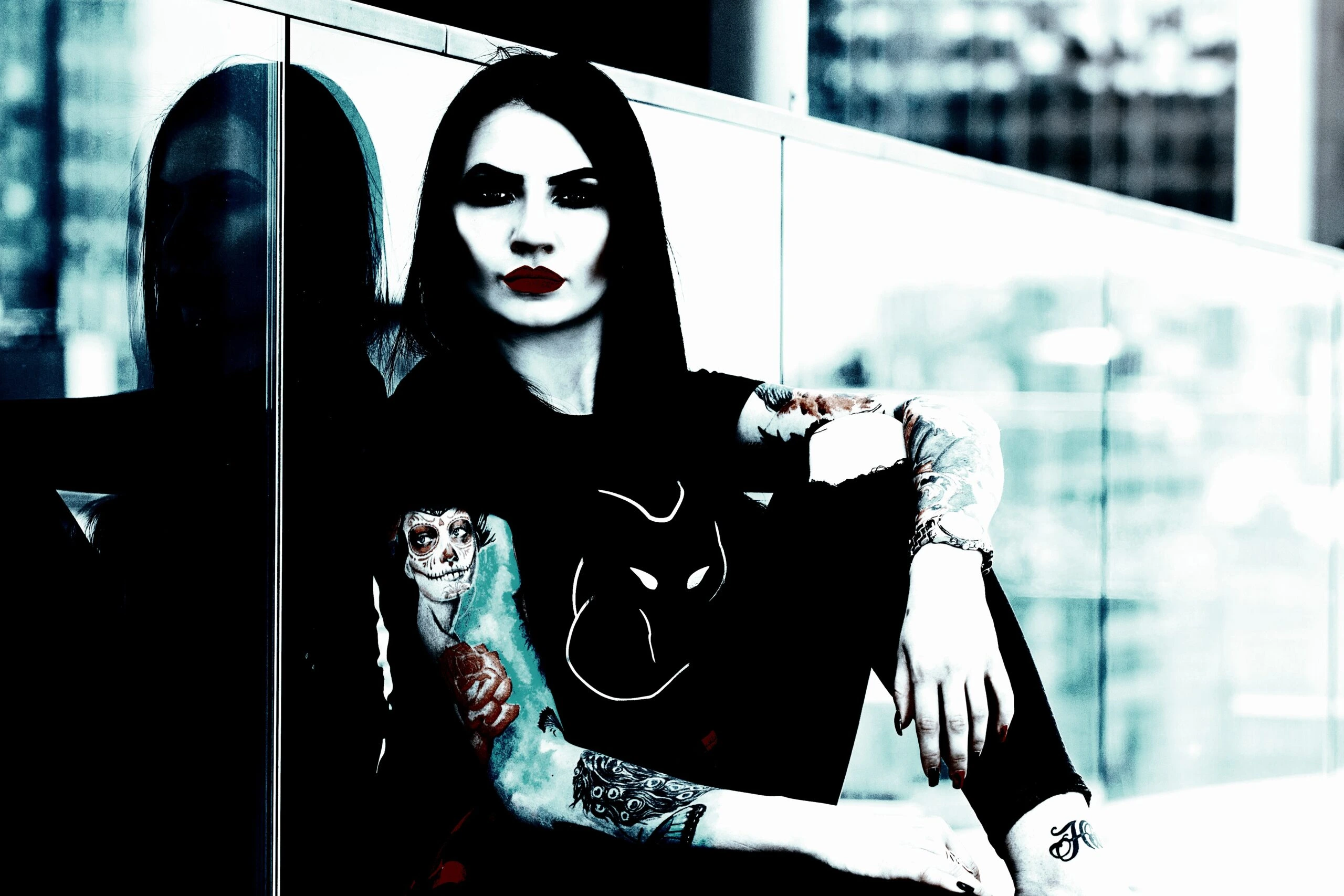 A gothic woman with dark hair and tattoos, wearing a black t-shirt with a white design, sitting in front of a reflective glass surface in an urban setting.