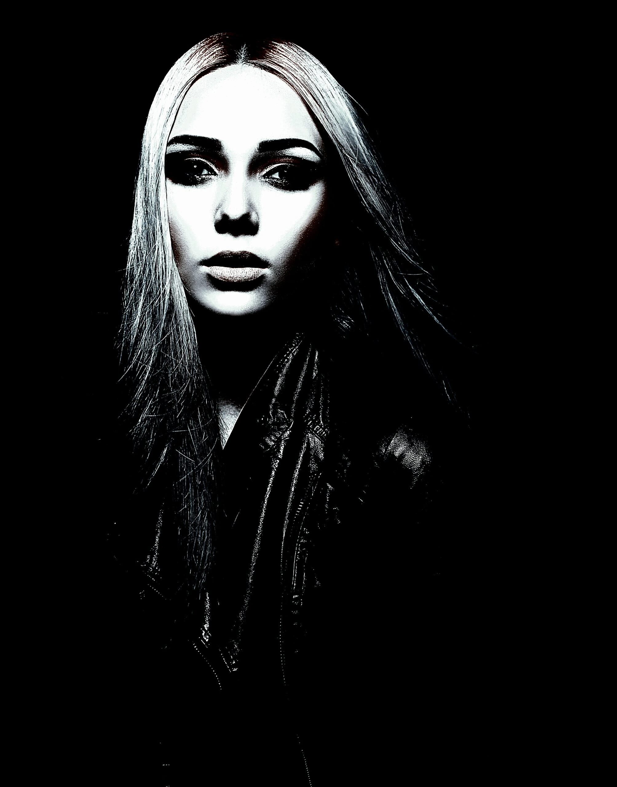 A gothic woman with pale skin and long dark hair, wearing dark makeup and a black leather jacket, against a black background.