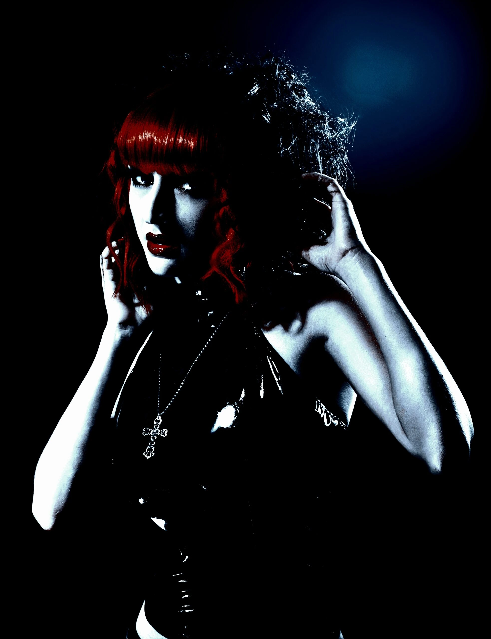 A gothic woman with red hair and a black outfit, accessorized with a cross necklace, against a dark background.