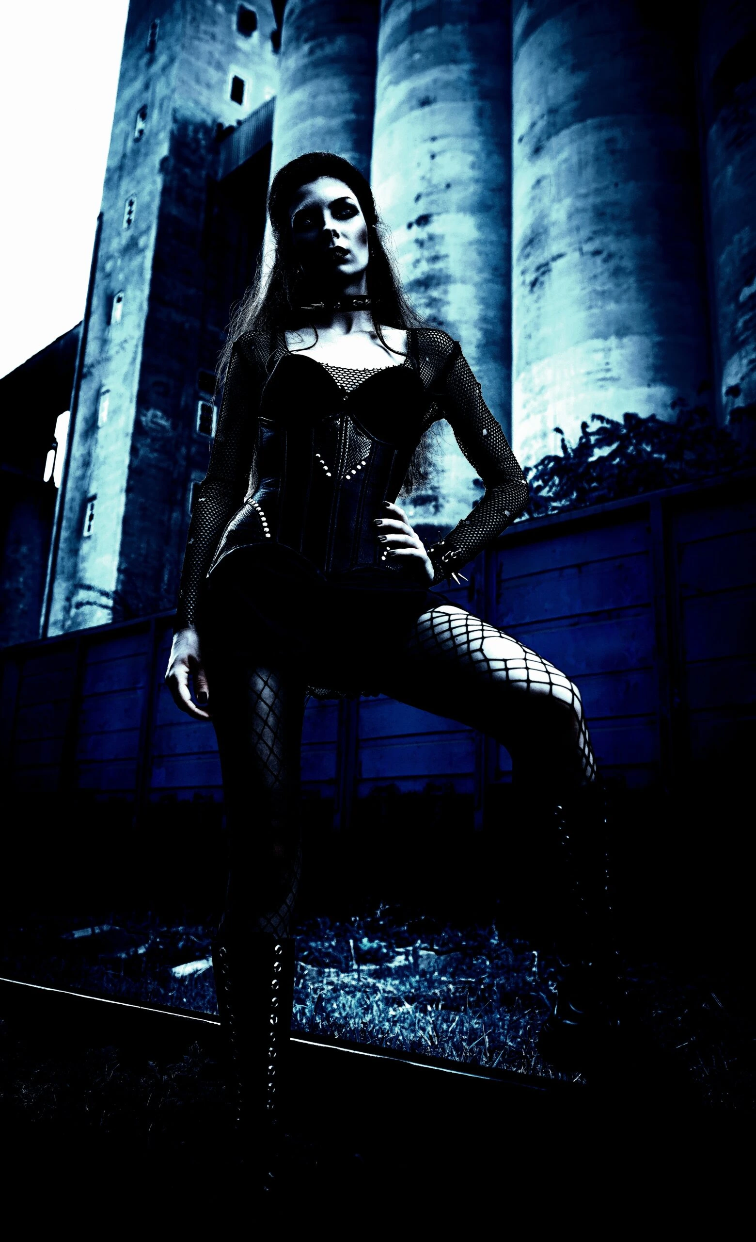 A gothic woman standing confidently in front of industrial silos, dressed in a black corset, fishnet sleeves, and fishnet stockings.
