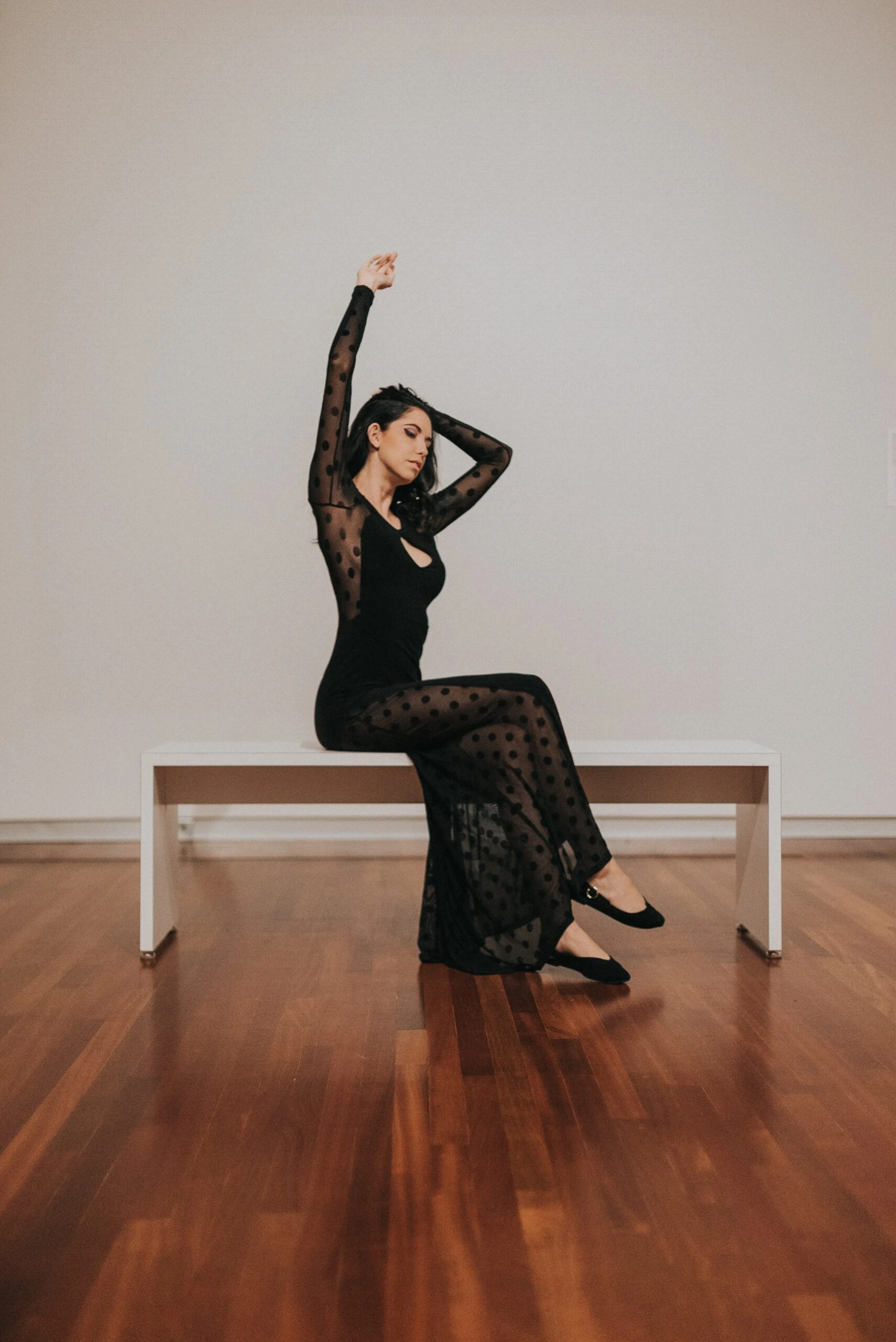 A woman in a gothic formal dress, featuring sheer polka dot fabric, striking a graceful pose with arms raised, seated on a white bench in a spacious room.