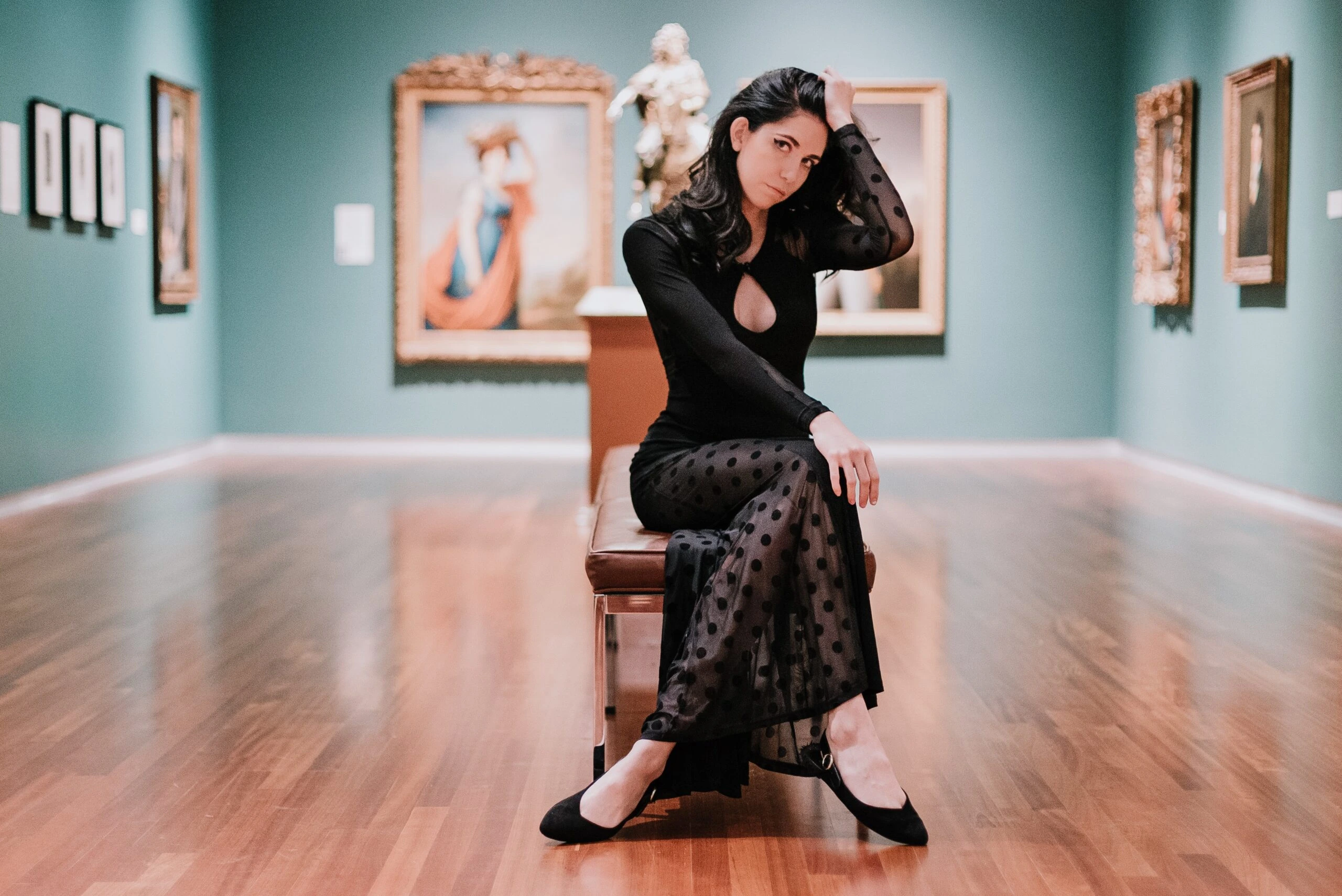 A woman wearing a gothic formal dress with sheer polka dot details, sitting thoughtfully on a bench in an art gallery surrounded by classic paintings.