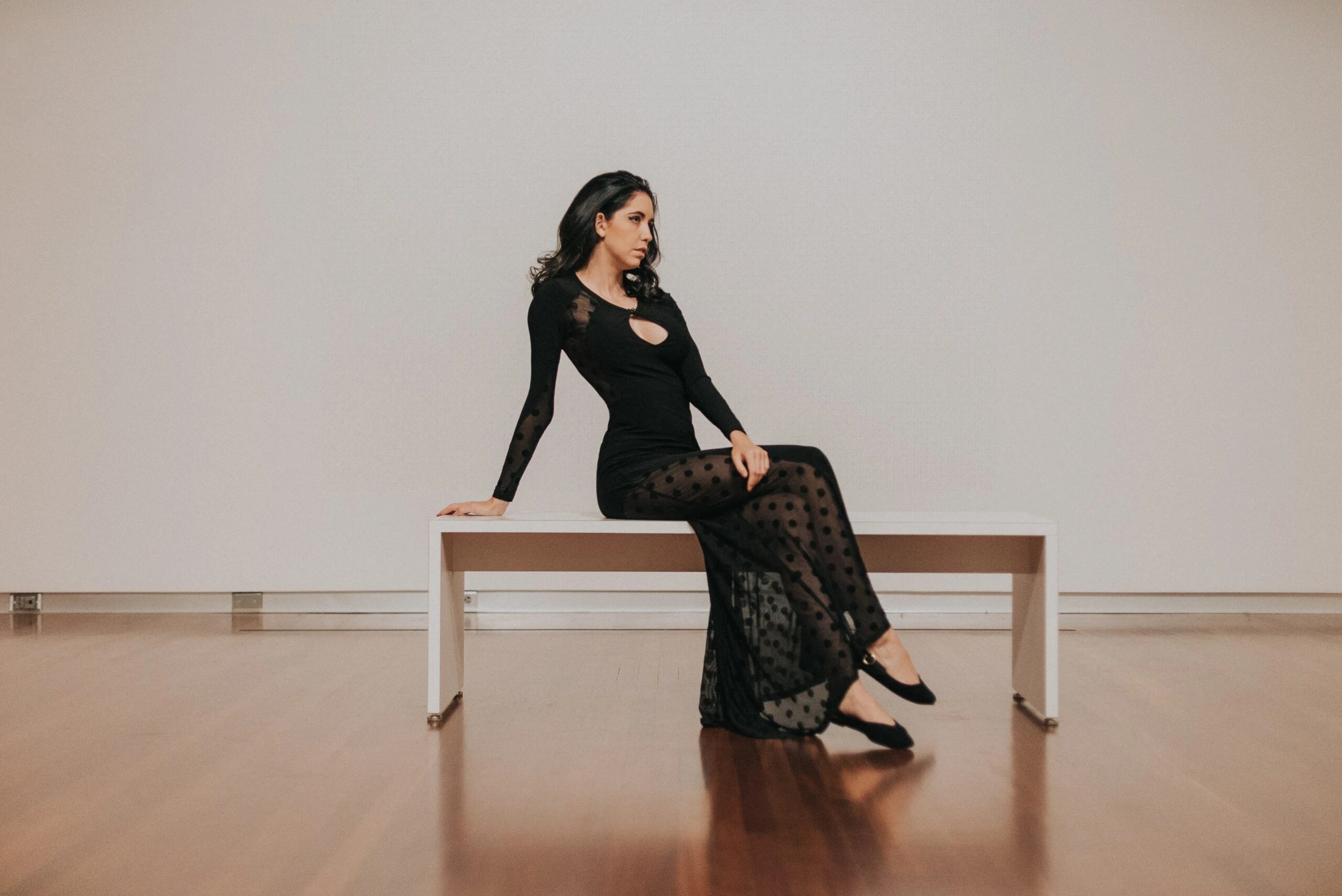 A woman in a sleek black gothic formal dress with sheer polka dot panels, sitting elegantly on a white bench in a minimalist room.