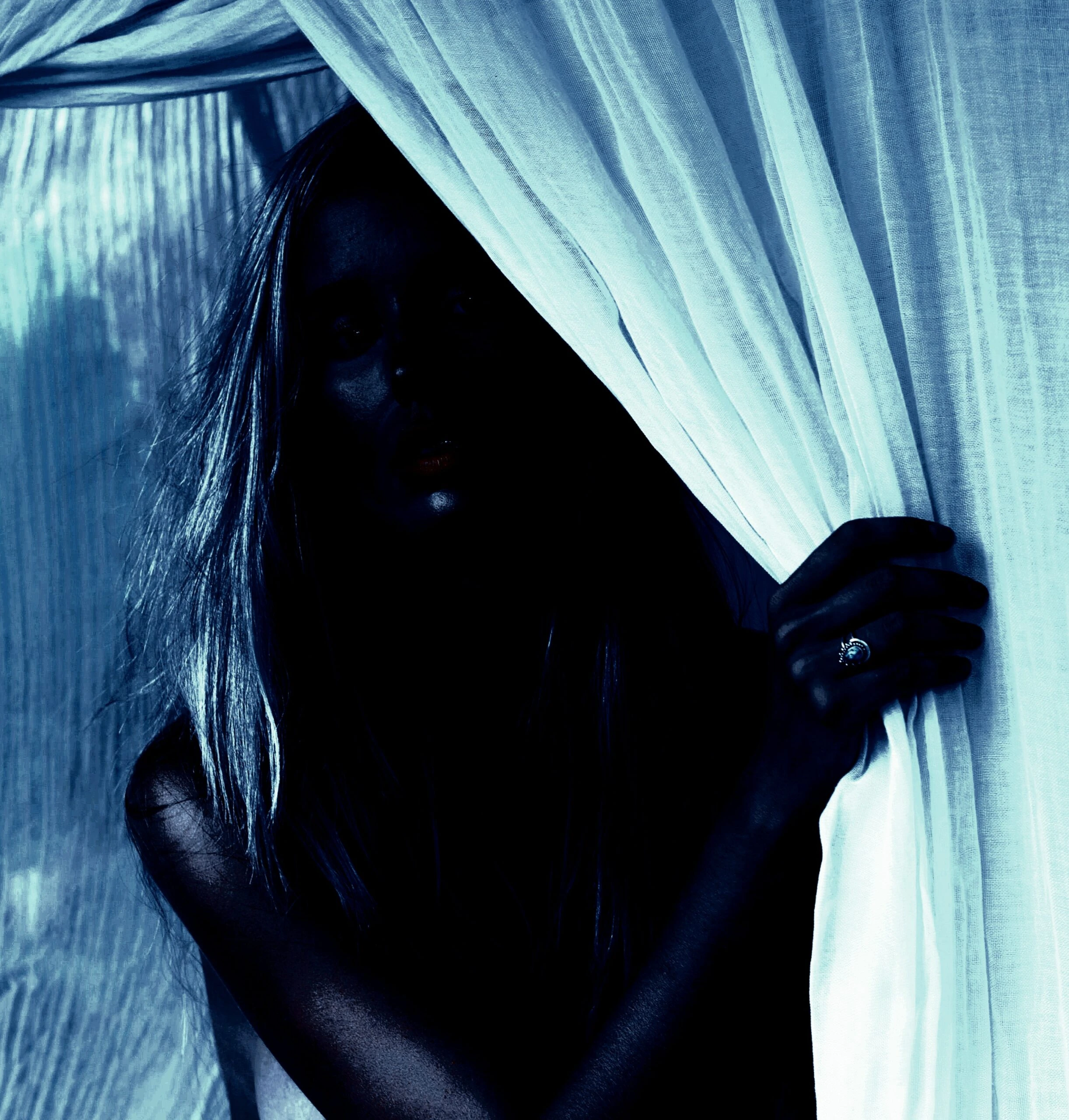 Woman in soft lighting with a mysterious expression, partially hidden behind curtains