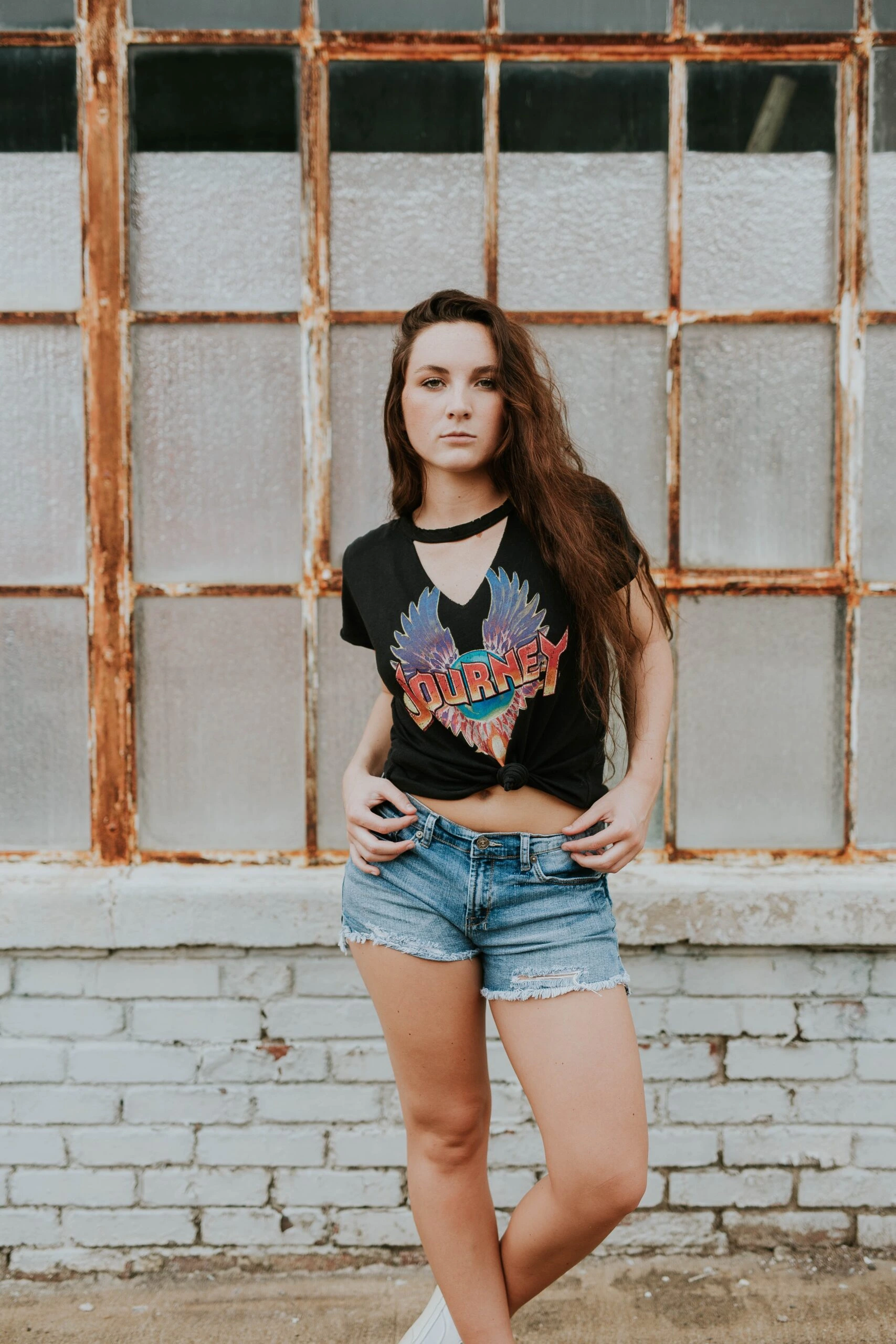 Woman in a Journey band T-shirt and denim shorts standing in front of a rustic window