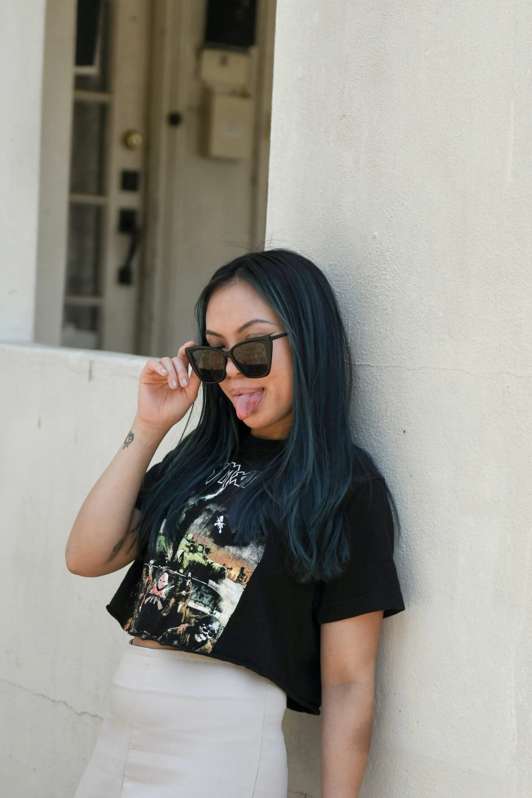 Woman with blue-tinted hair wearing a black Iron Maiden band T-shirt and sunglasses posing against a white wall