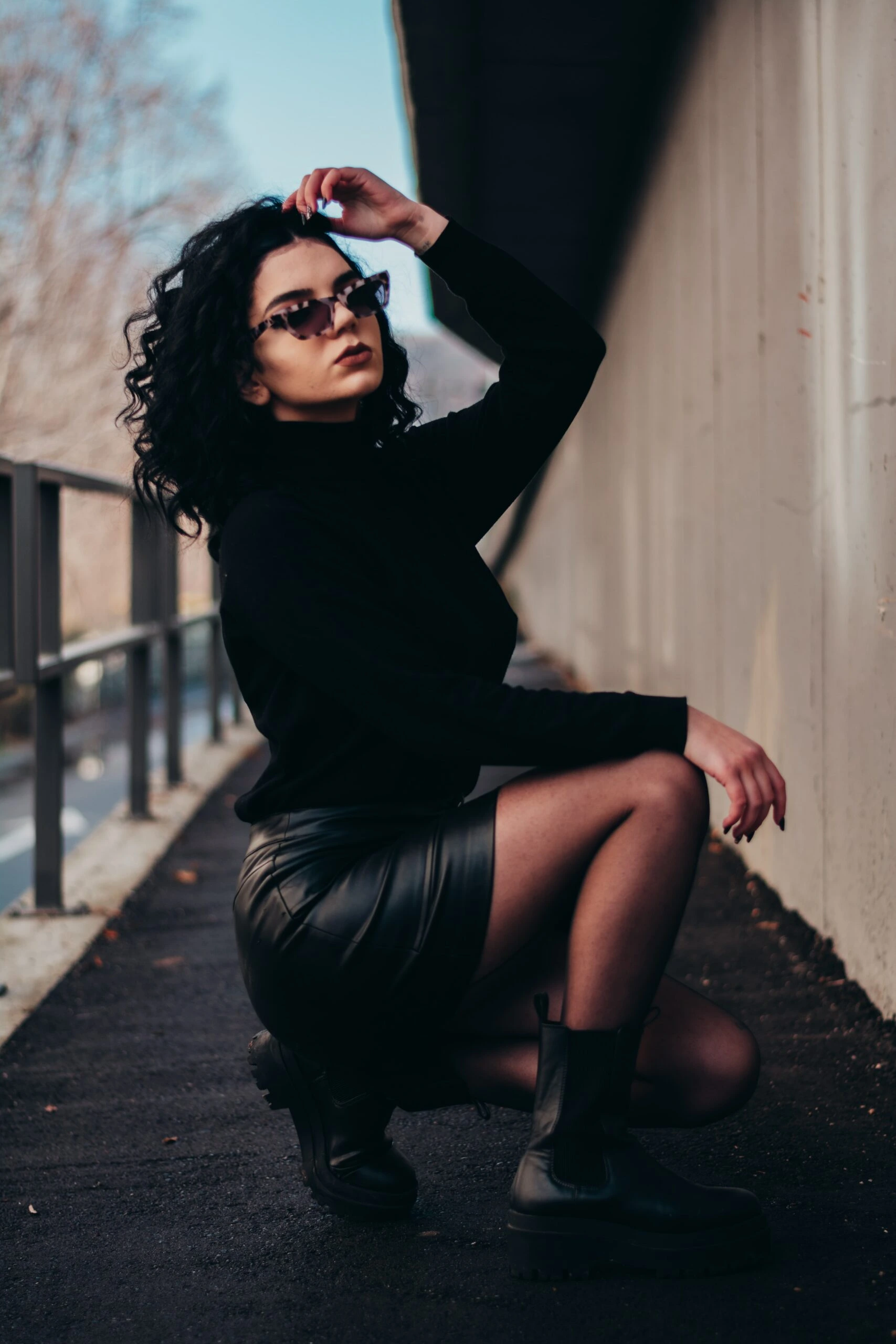 Stylish woman in black attire and sunglasses, kneeling on a city street, representing the allure of wearing sunglasses at night.